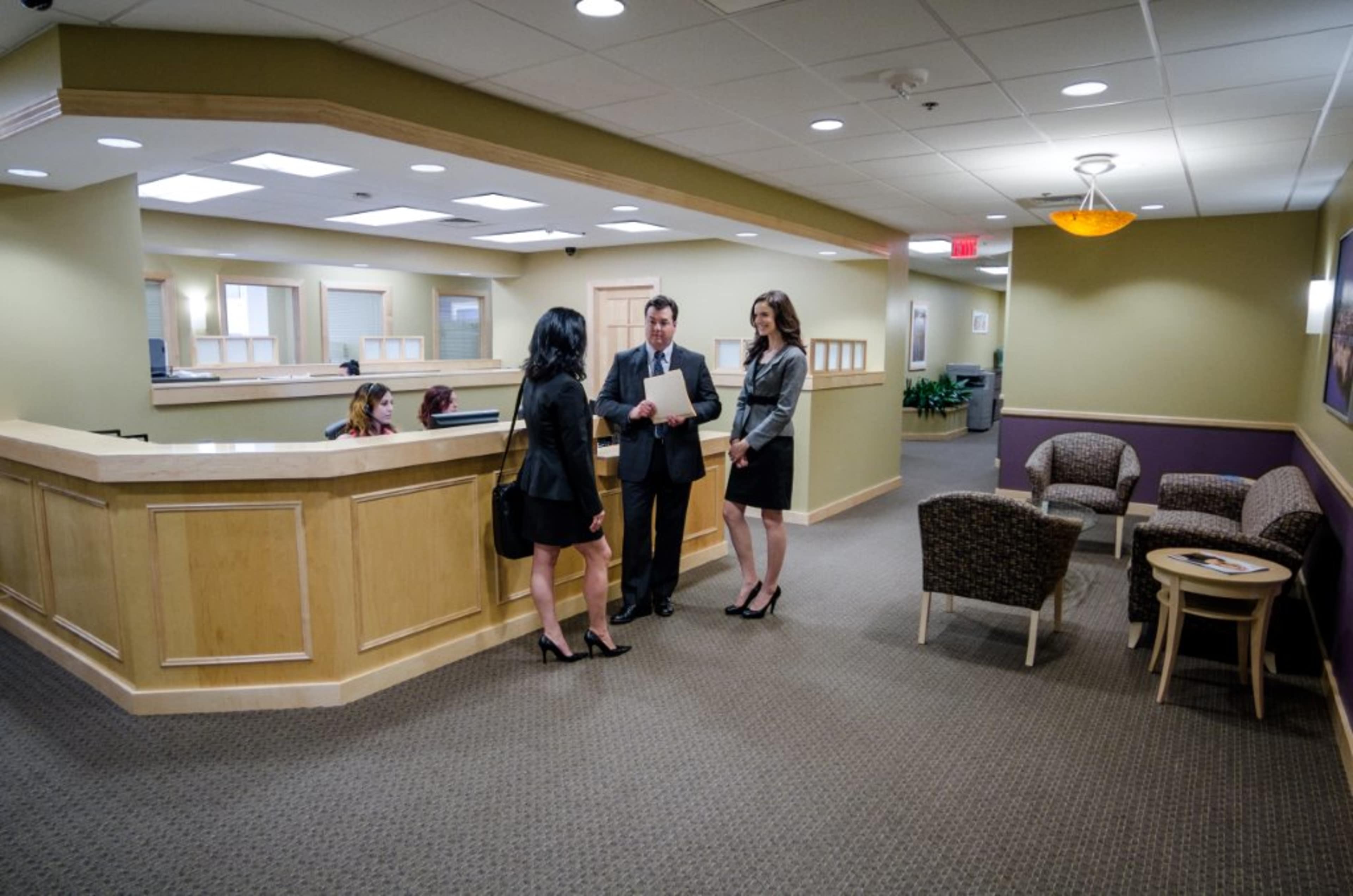 Three professionals in formal attire are discussing near a reception area in an office setting, while two individuals are seated in the background.