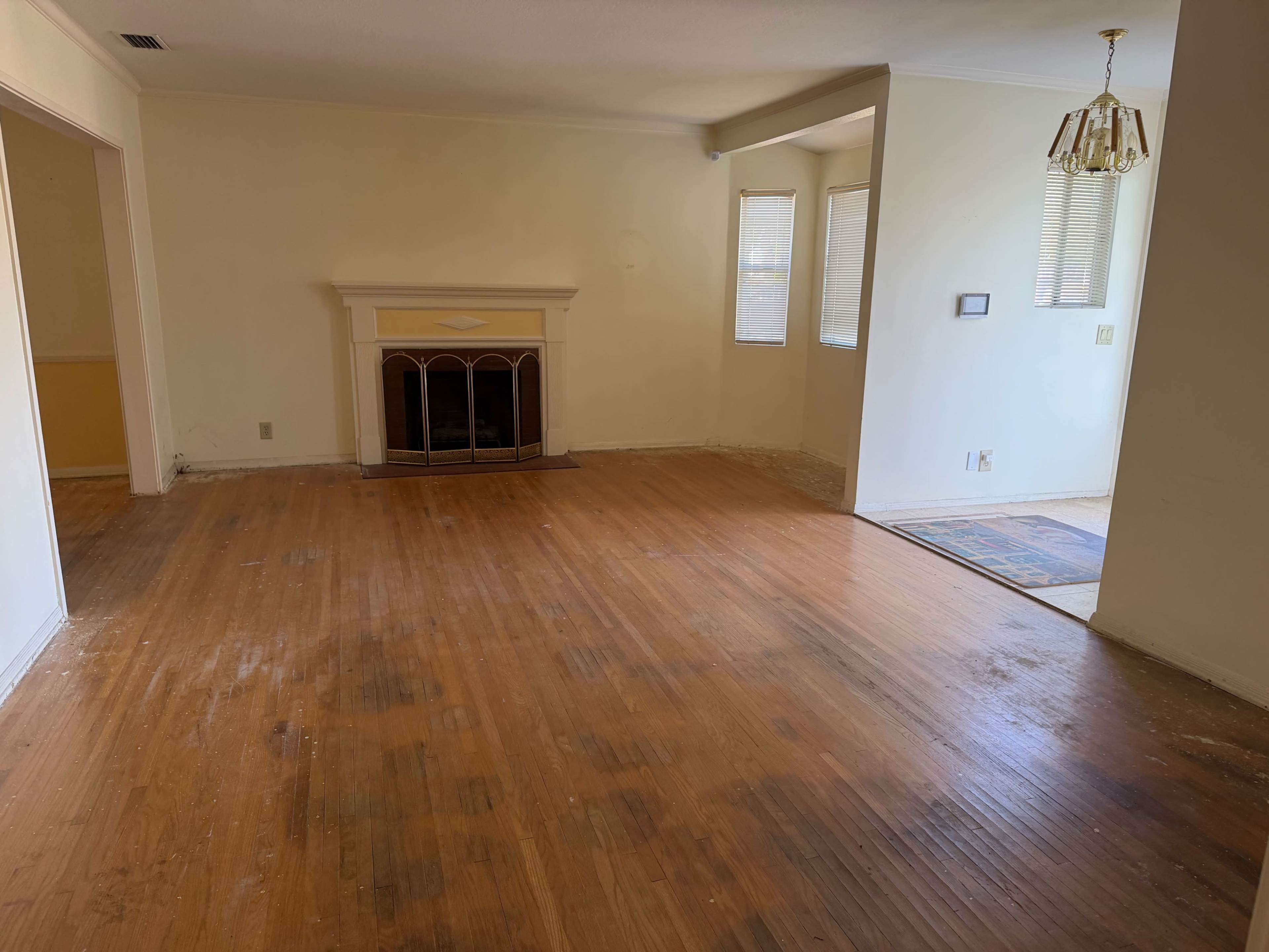 The image shows an empty living room with wooden floors, a fireplace in the center, and a ceiling light fixture.