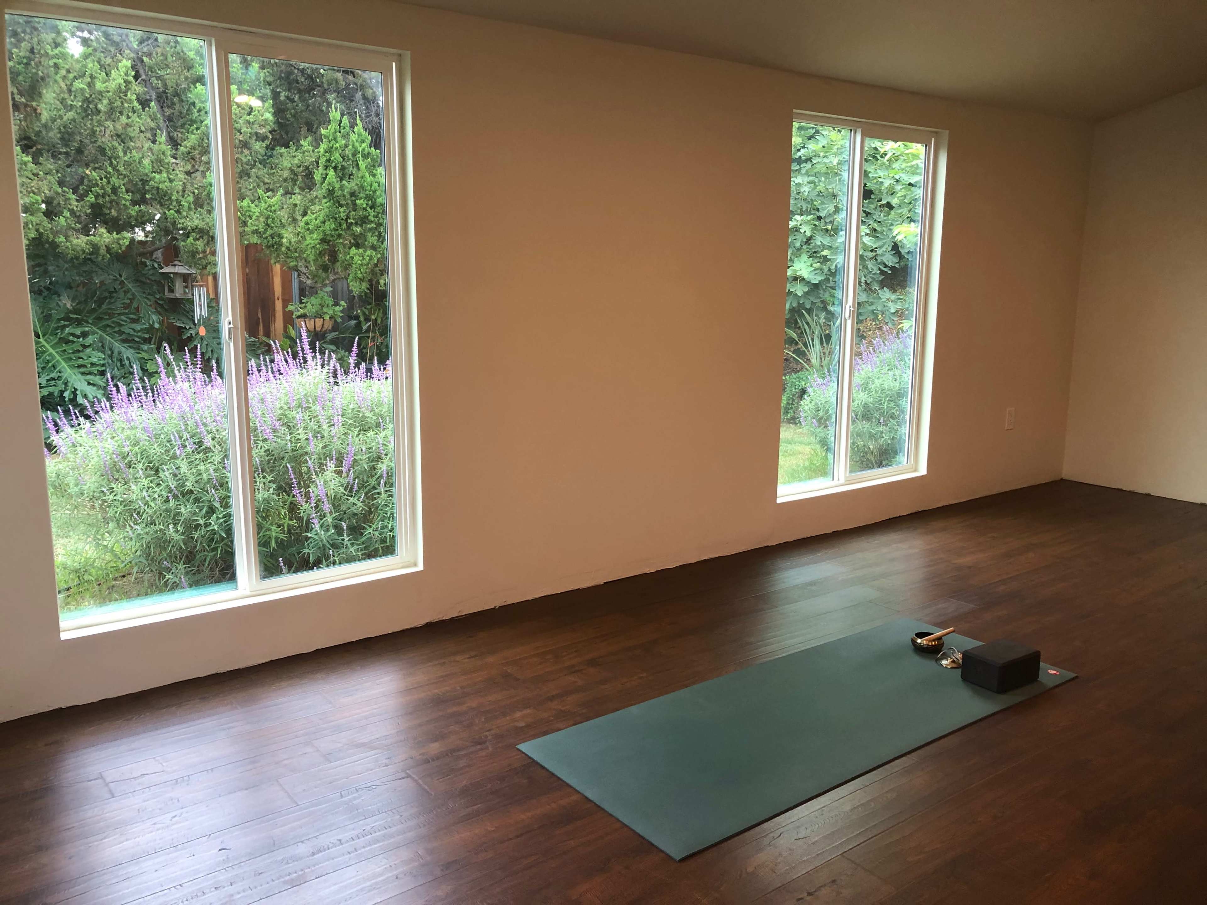 A yoga mat with props is placed on a wooden floor in a room featuring large windows overlooking a garden.