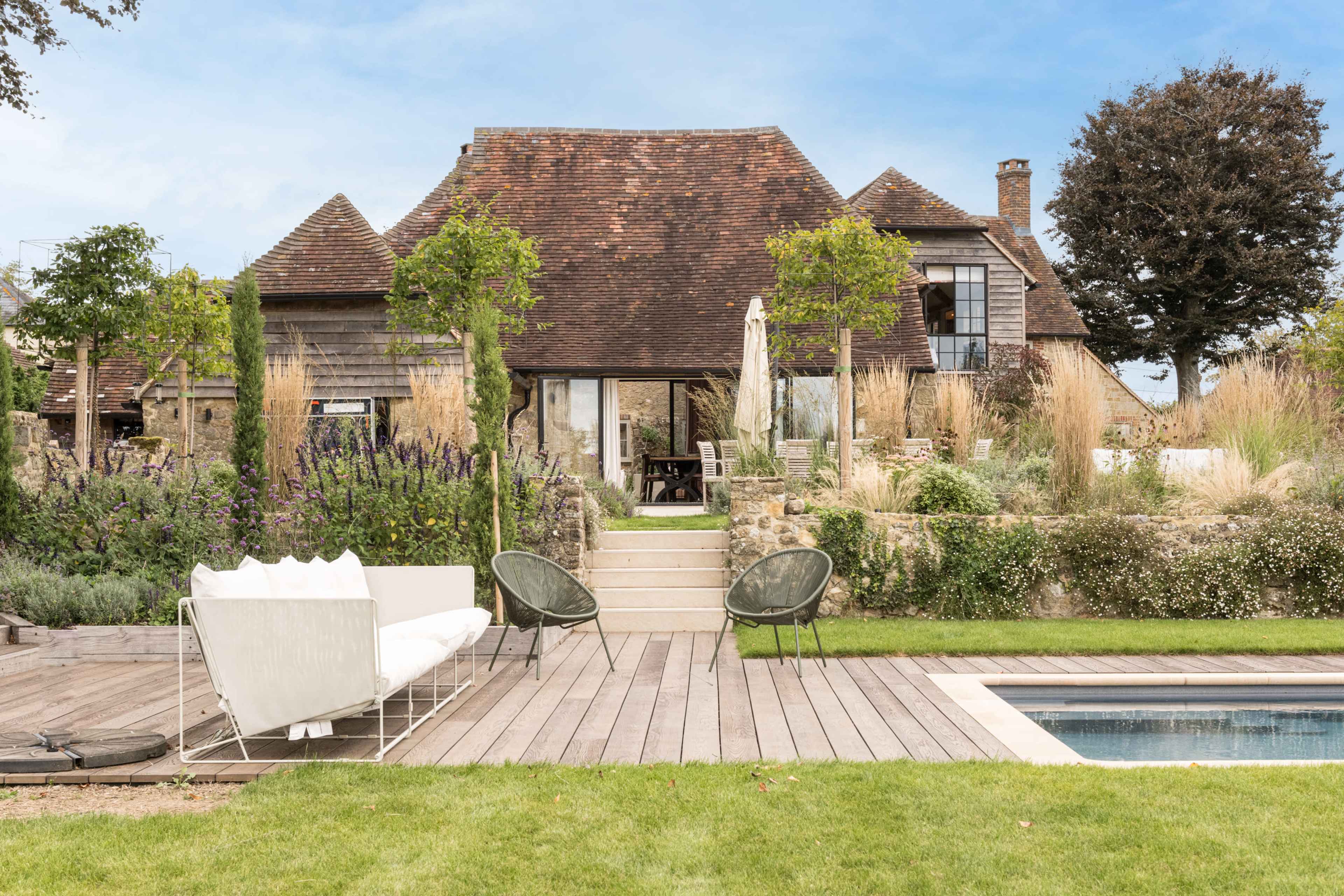 A modern house with a steeply pitched roof is situated behind a landscaped garden featuring steps, a wooden deck, and a swimming pool.