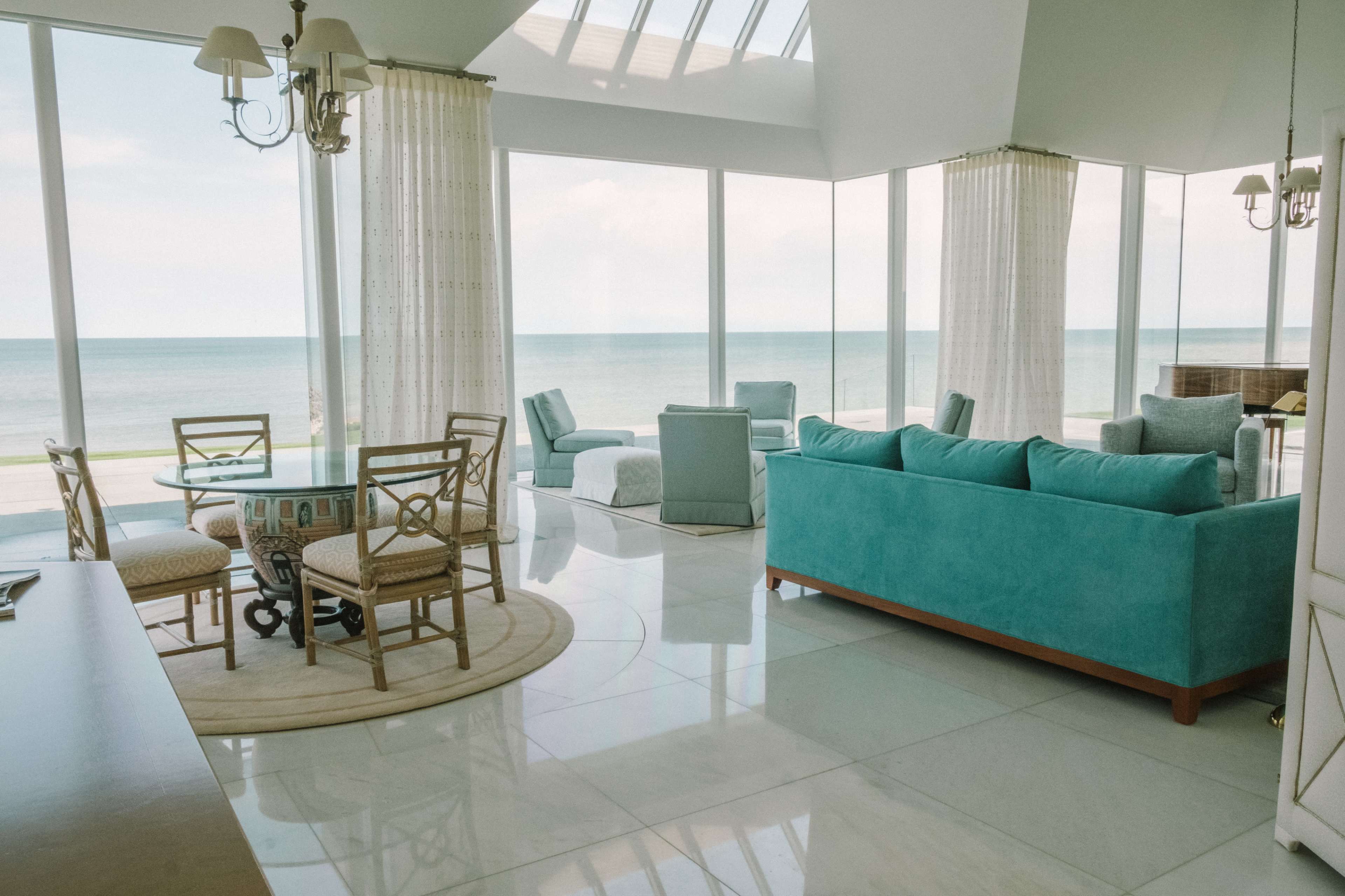 The image shows a modern living room with large windows overlooking the ocean, featuring turquoise furniture, a round dining table, and a beige rug on a tiled floor.