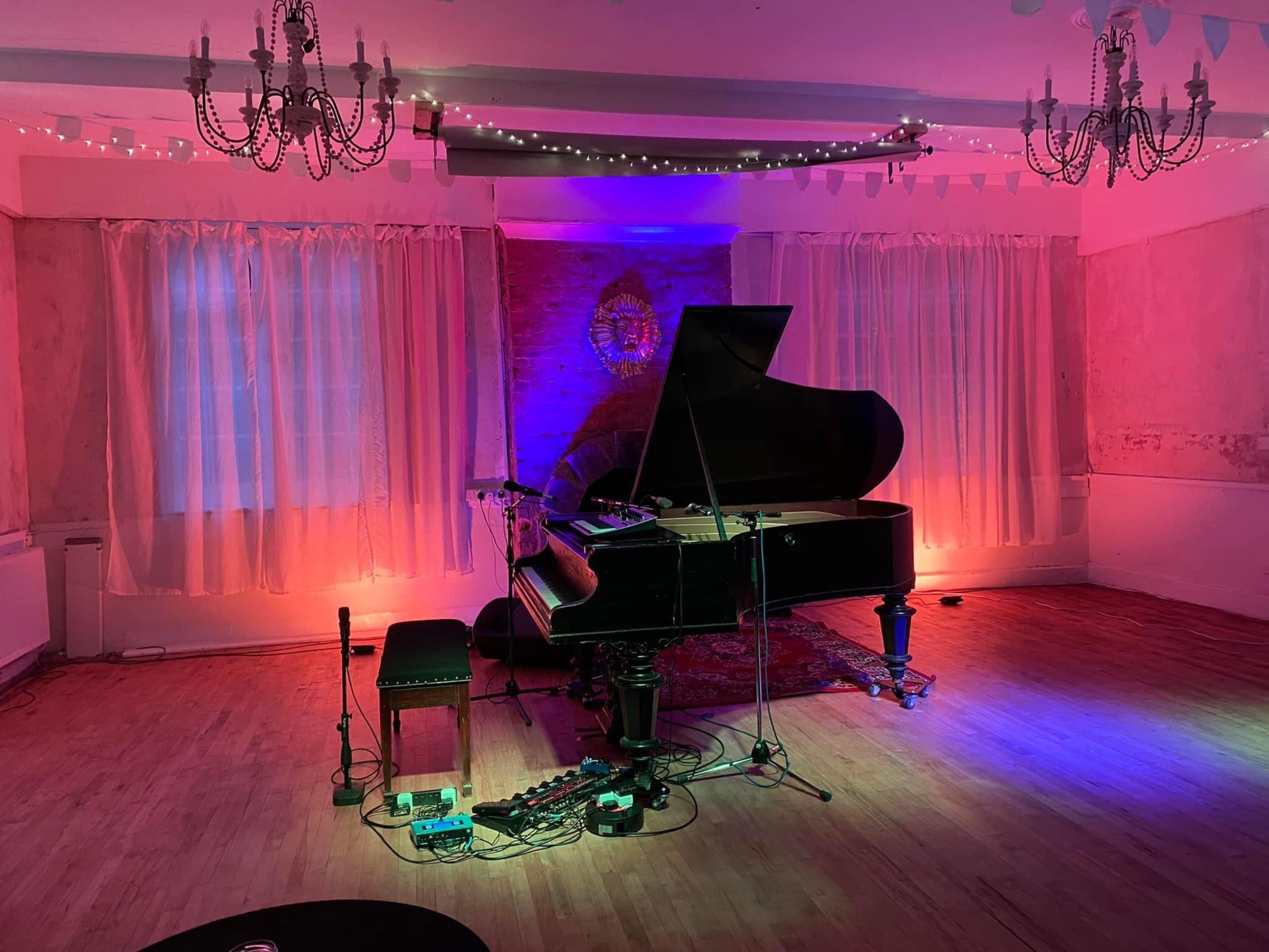 A grand piano sits in a sparsely decorated room with colorful lighting, surrounded by musical equipment and a microphone stand.