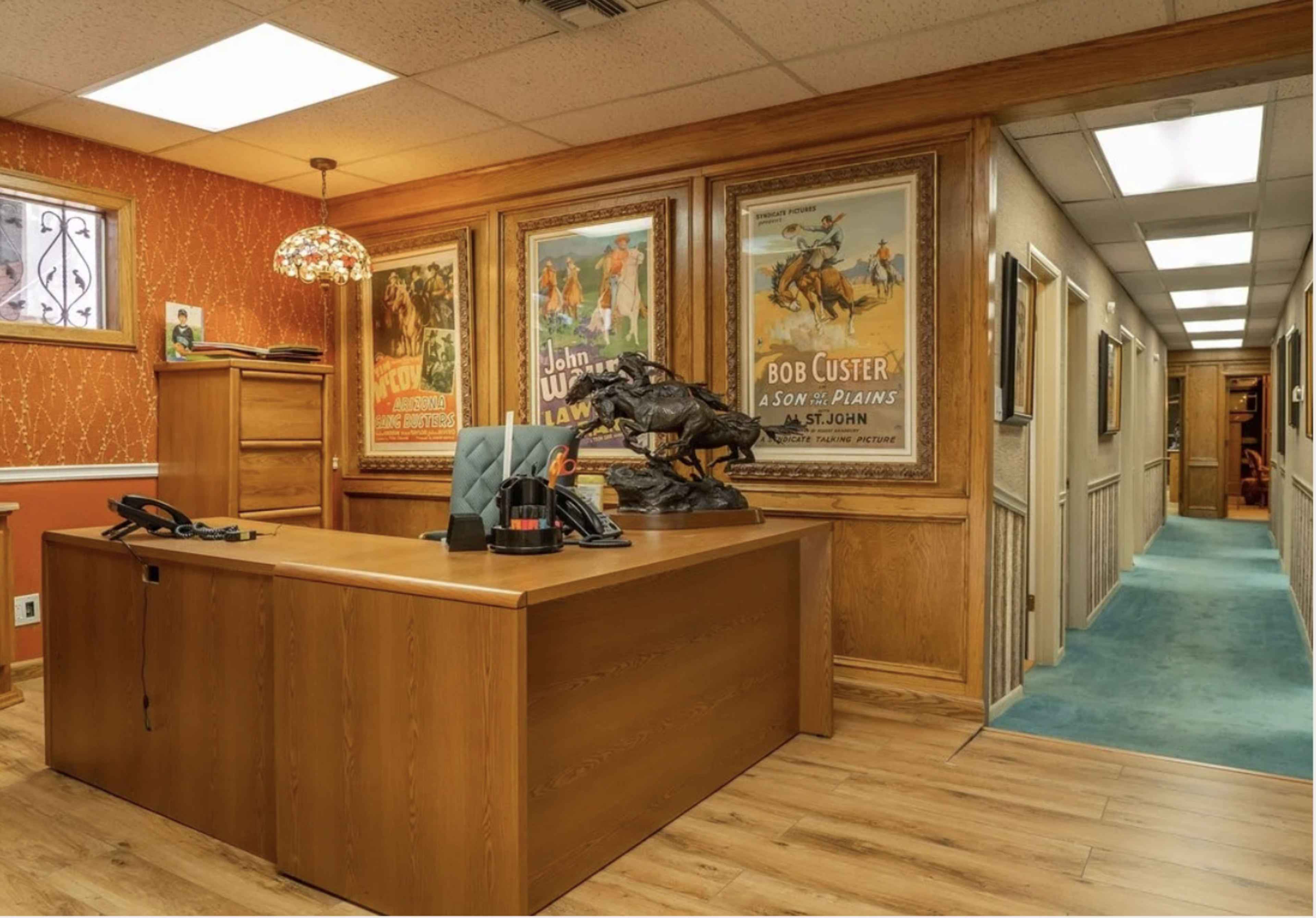 The image shows an office space featuring wooden furniture, mounted Western-themed posters on the wall, and a hallway leading to another part of the building.