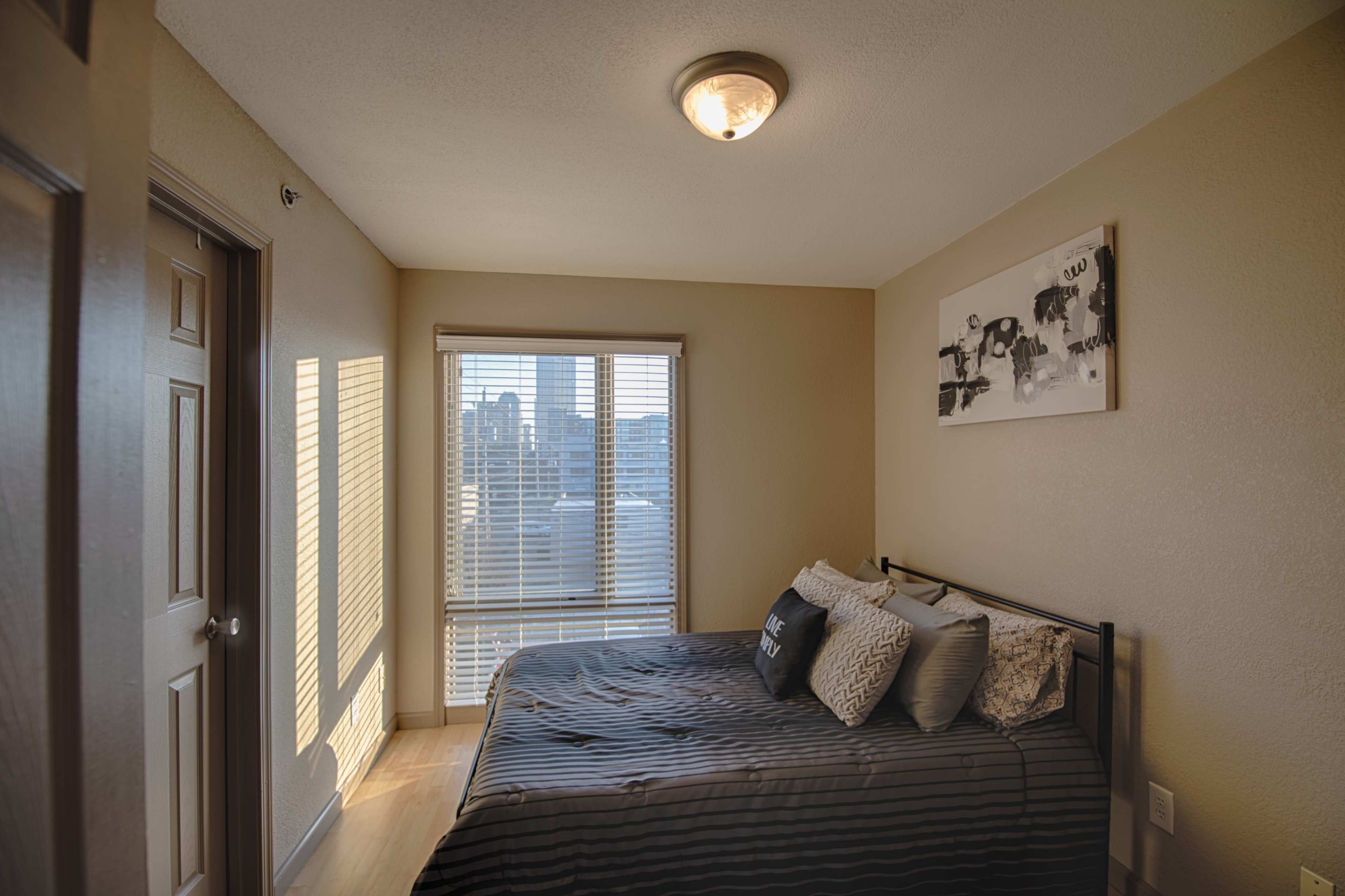 A small bedroom with a bed, a window featuring blinds, and a piece of artwork on the wall.