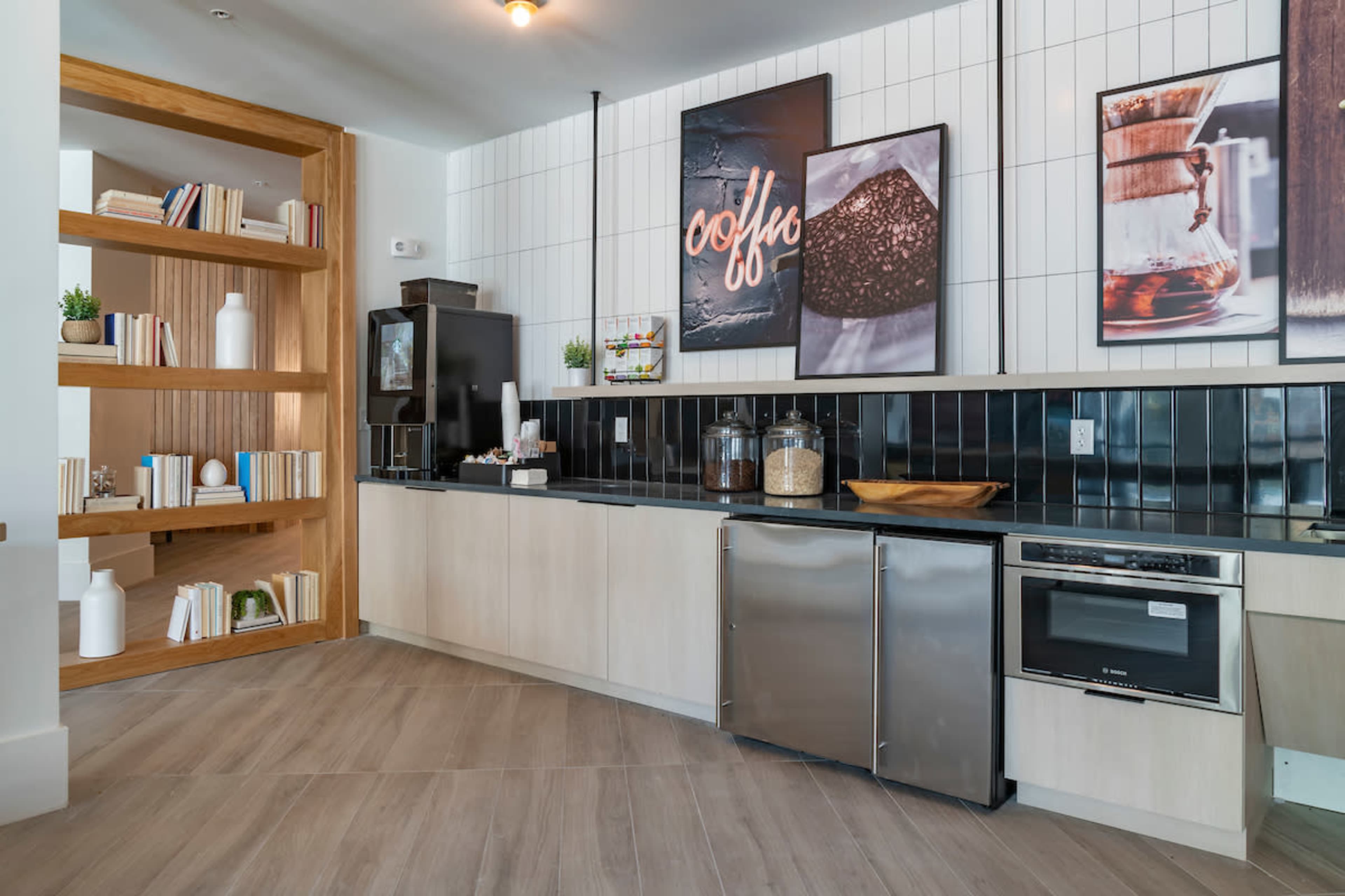 A modern kitchen area featuring a black and white tiled backsplash, wooden shelves with books, and various coffee-related artwork on the walls.