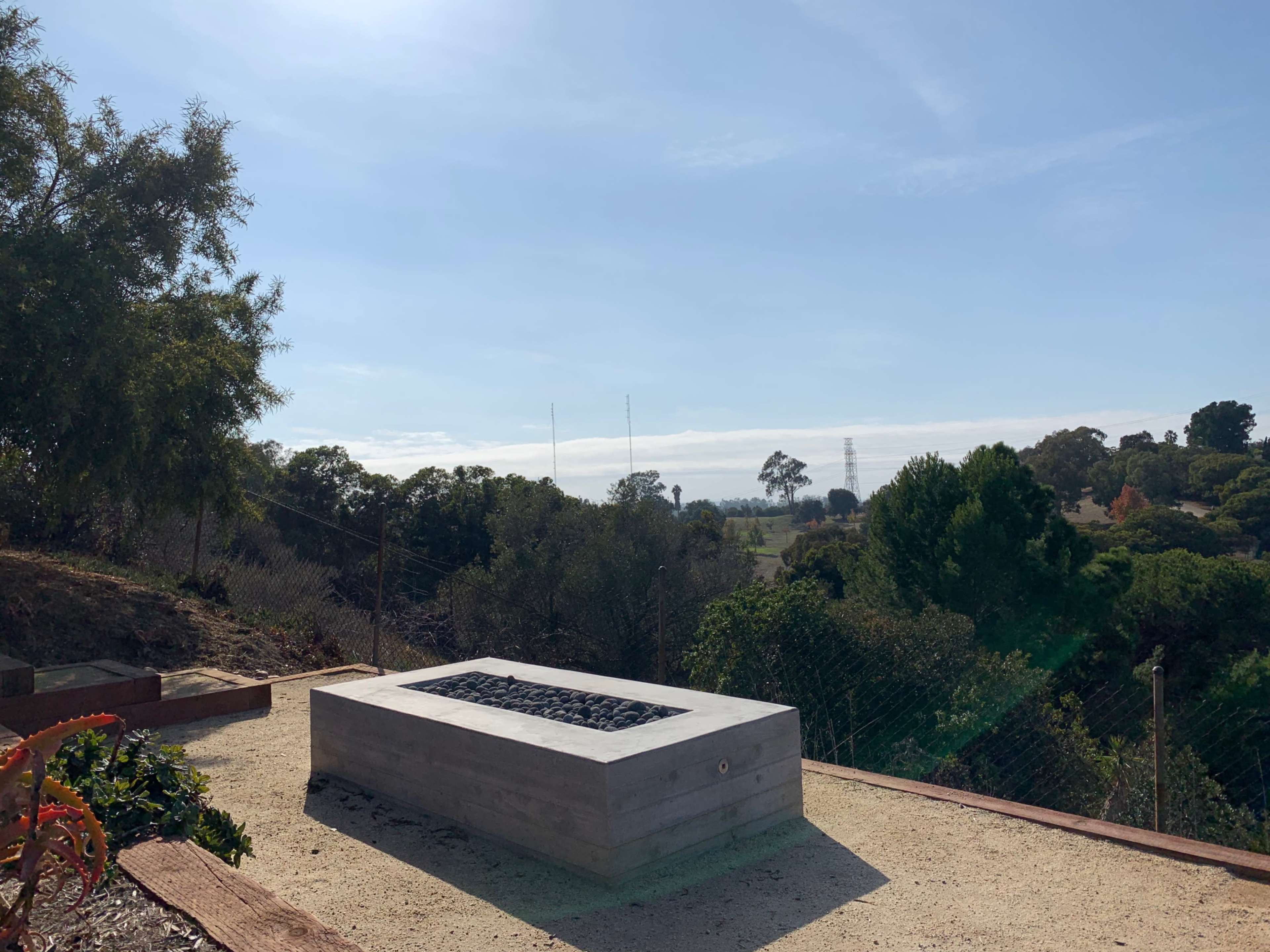 A rectangular concrete fire pit set on a gravel surface, surrounded by trees and brush under a clear sky.