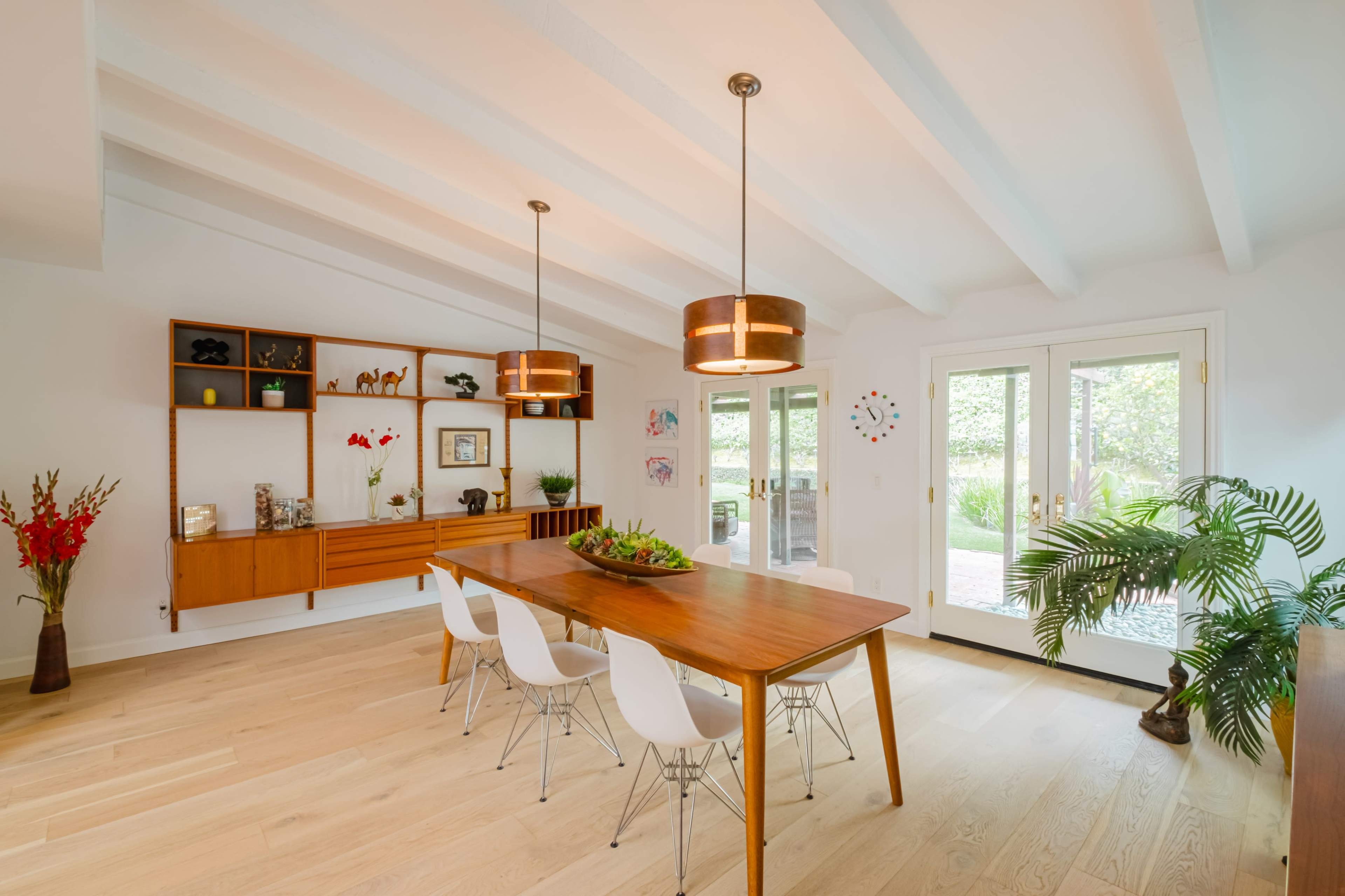 A modern dining room featuring a wooden table with white chairs, a sideboard, and large glass doors opening to an outdoor space.