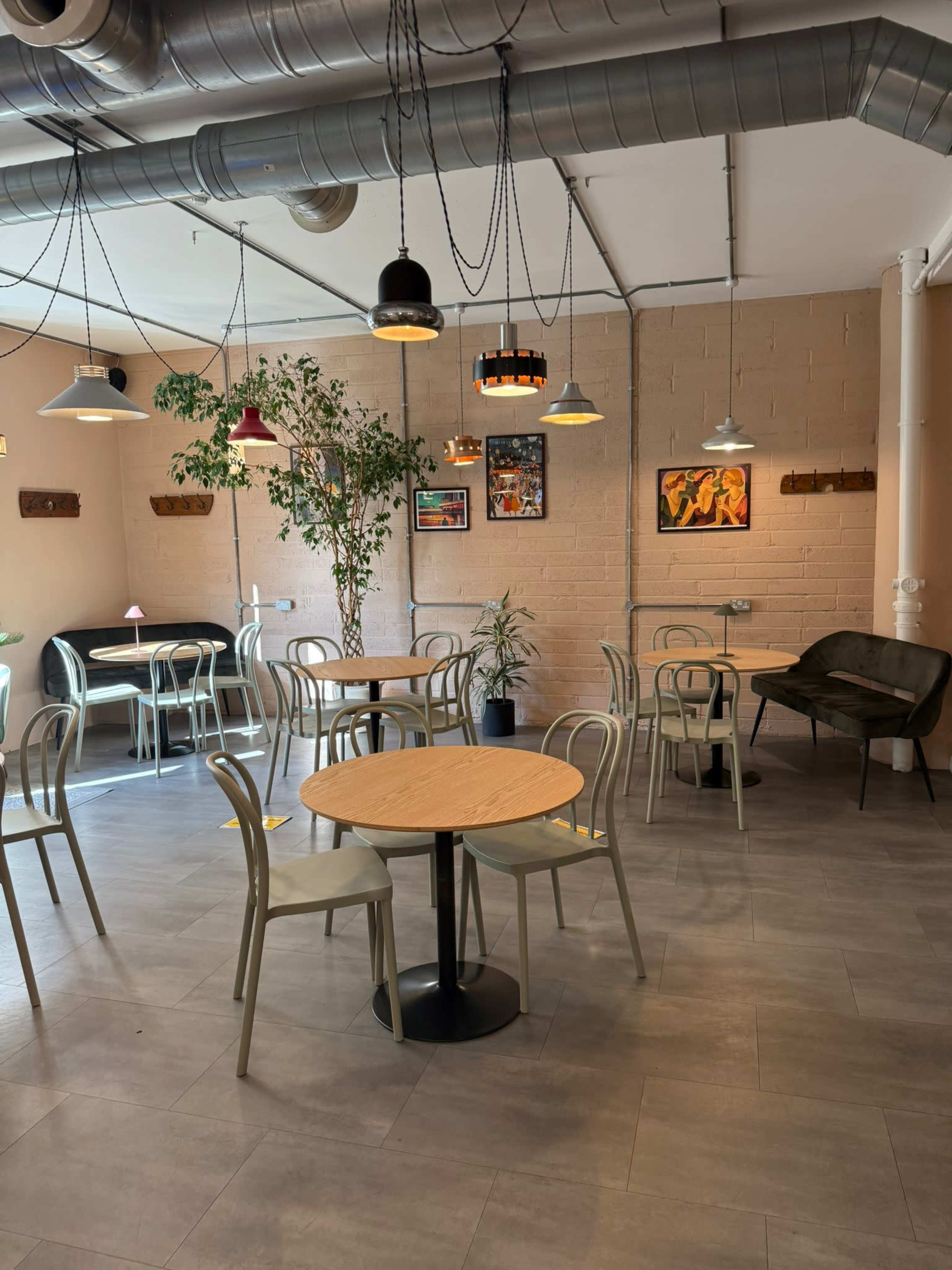 The image shows a modern café interior with several round tables and chairs, artwork on the walls, and various pendant lights hanging from the ceiling.