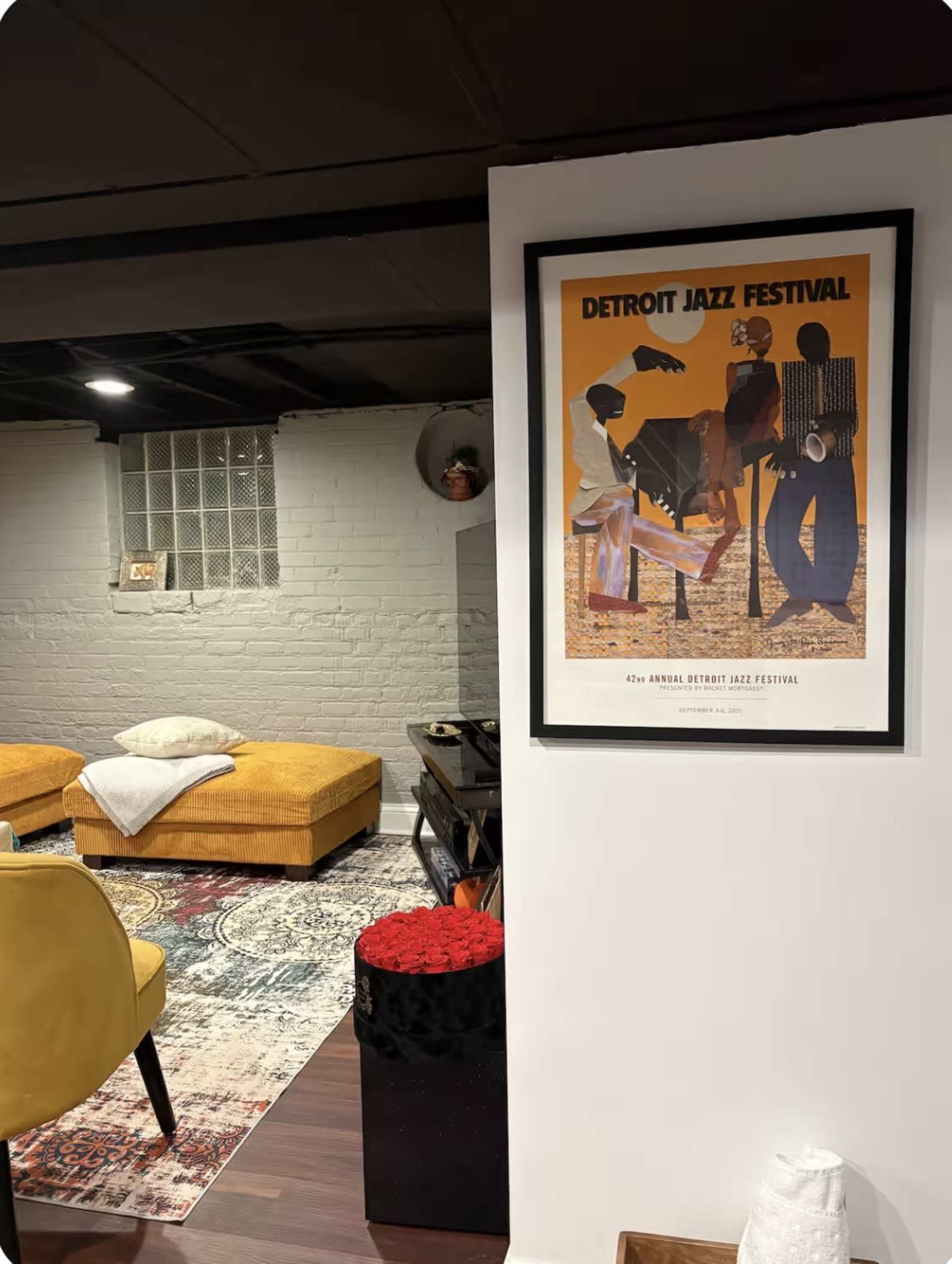 The image shows a basement area with a framed poster for the Detroit Jazz Festival on the wall, featuring a colorful design, and a modern seating arrangement with yellow ottomans and a patterned rug.