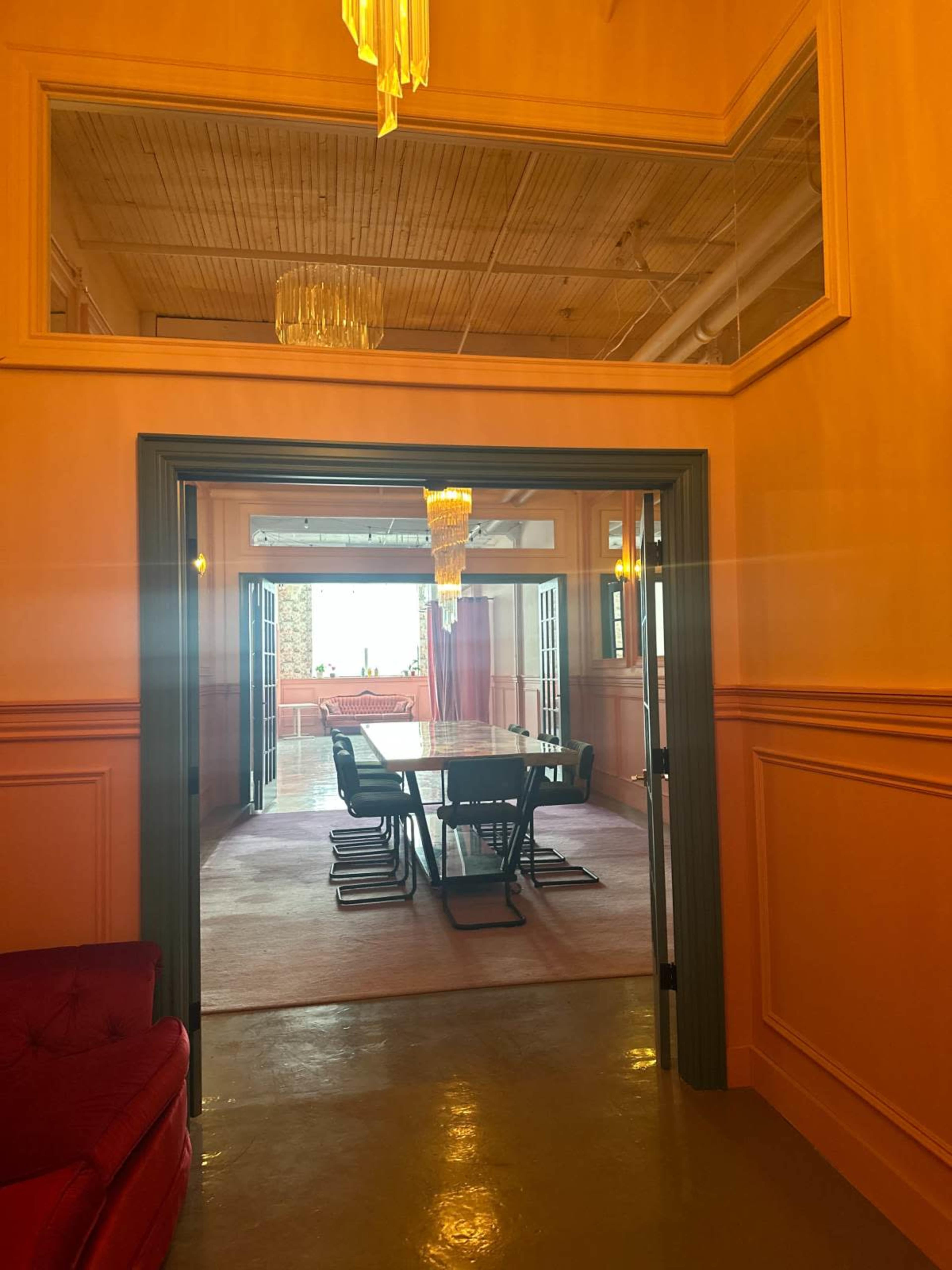 A hallway leads to a well-lit dining area with a large table, floor-to-ceiling windows, and a chandelier.