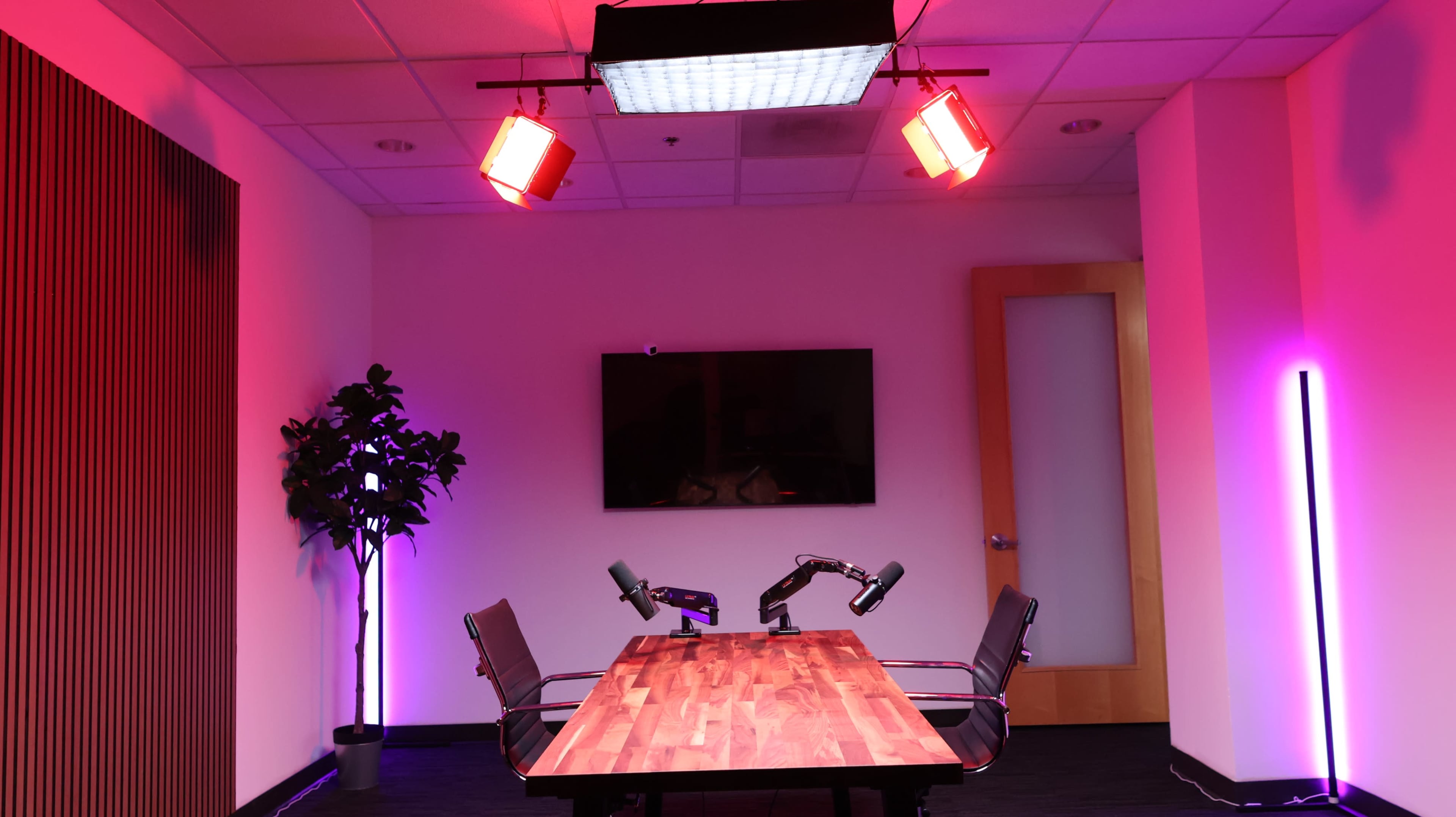 A modern conference room with a wooden table, two microphone arms, a large wall-mounted screen, and vibrant colored lighting.