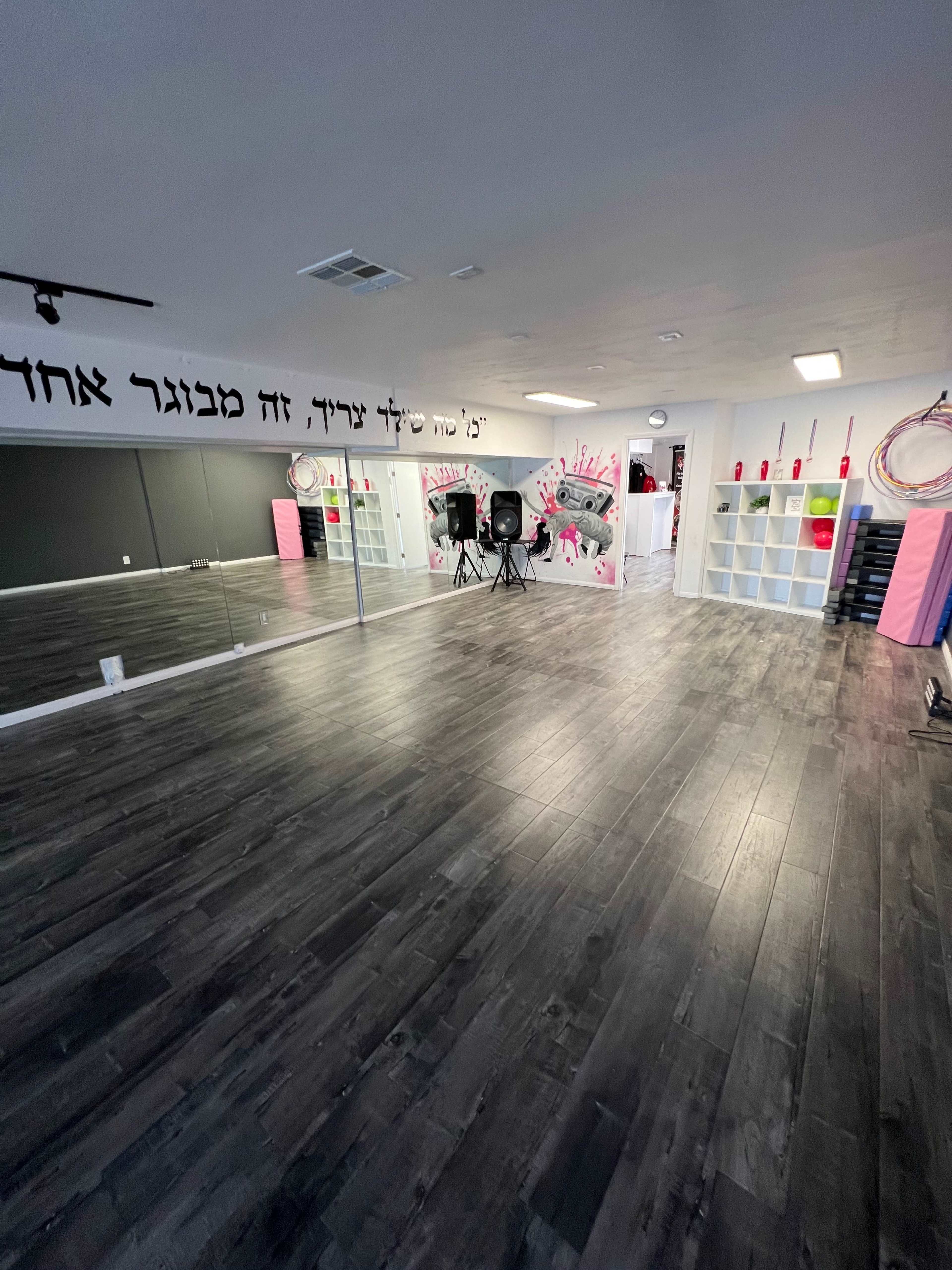 An empty dance studio with a mirrored wall, black flooring, and storage shelves adorned with pink and white accents.