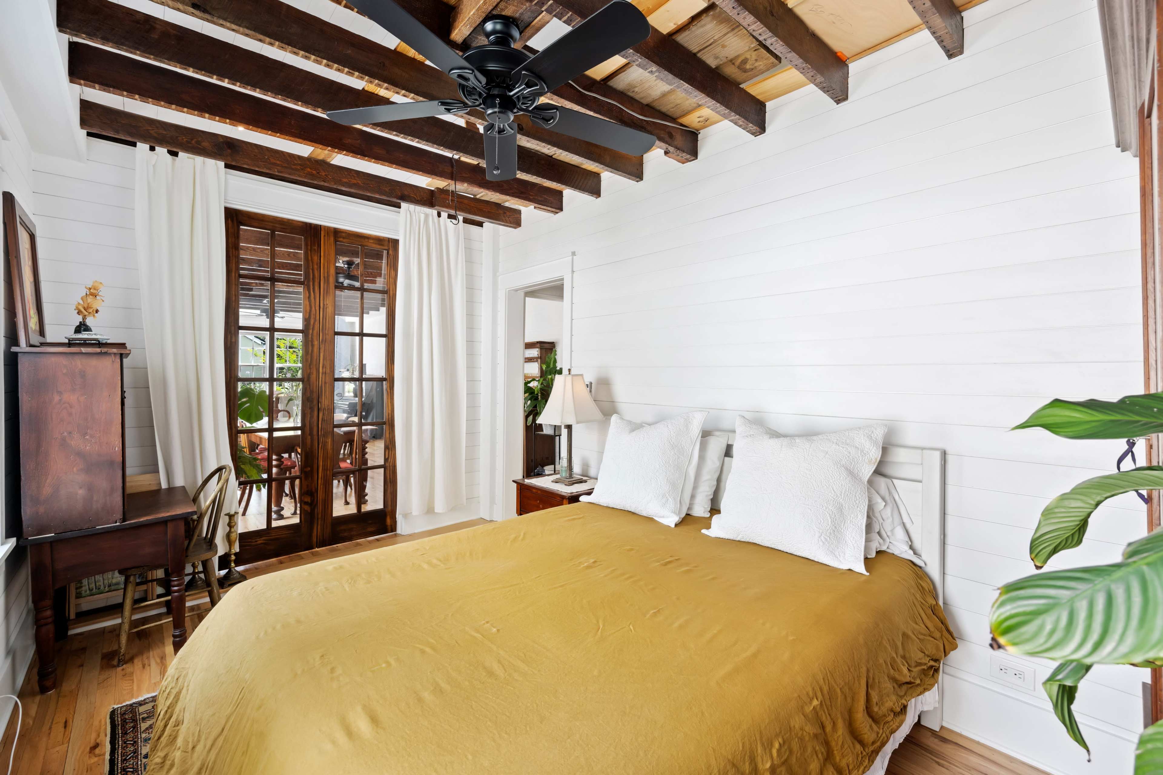 The image shows a well-lit bedroom with a yellow bedspread, wooden beams on the ceiling, and a view of a patio through double doors.