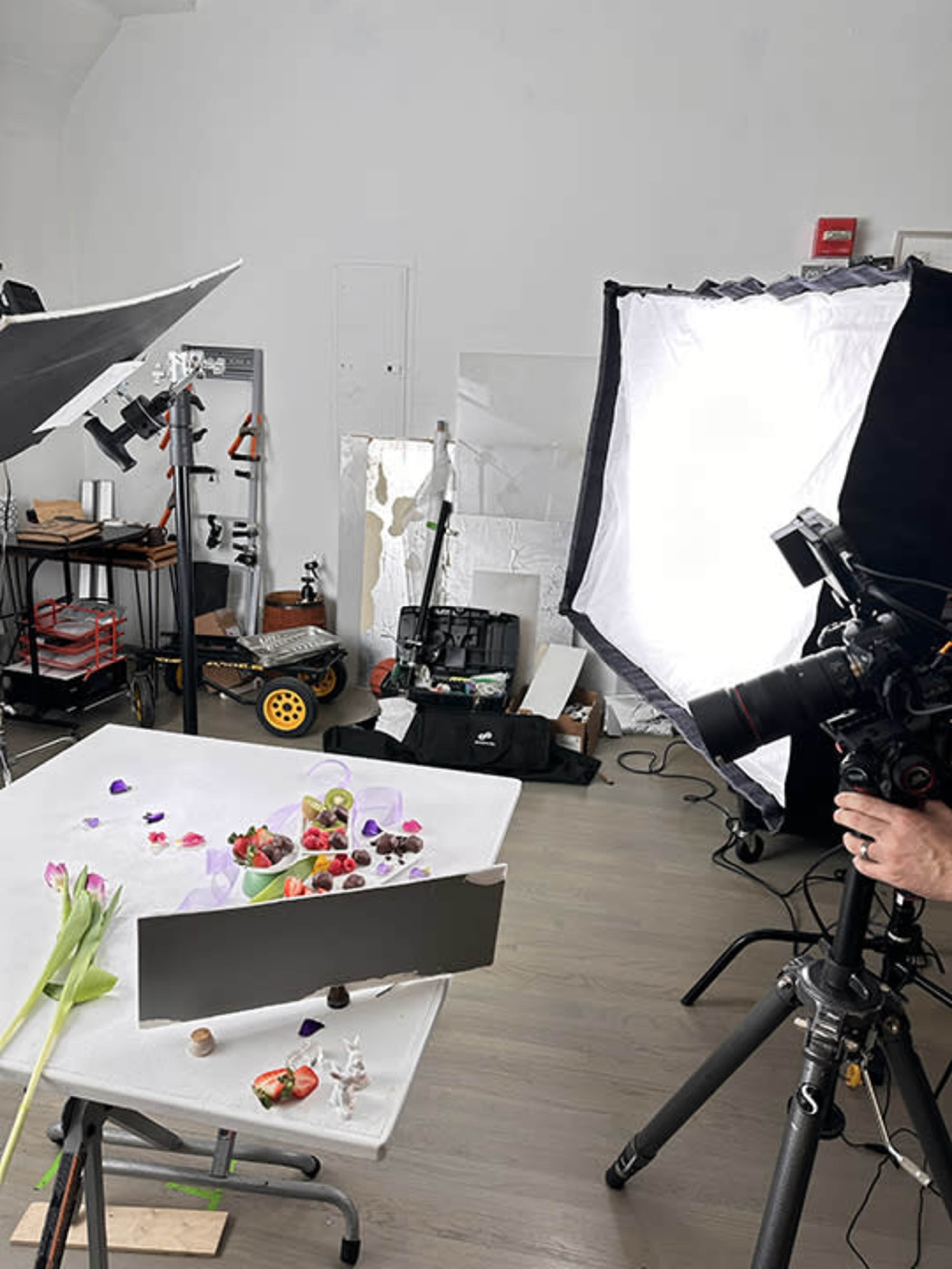 The image shows a photography studio setup with a table displaying various food items, camera equipment, and lighting tools.