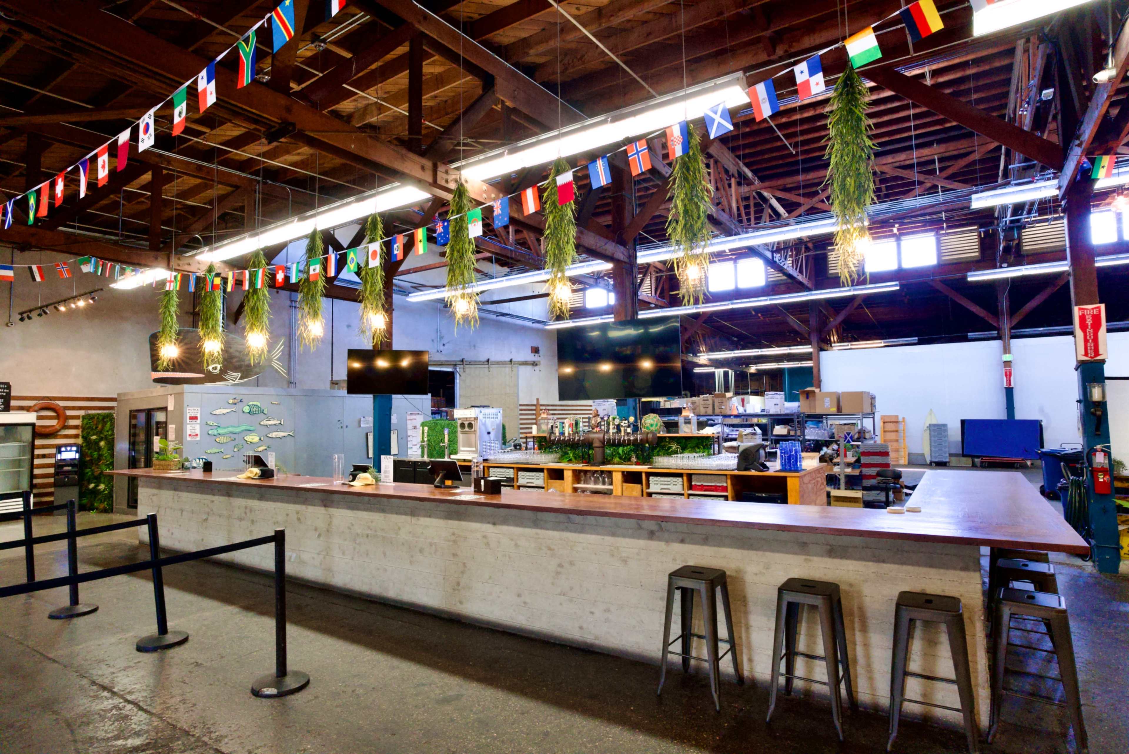 The image shows a modern bar area with a long wooden counter, hanging greenery, and flags from various countries adorning the ceiling in a spacious, industrial-style interior.