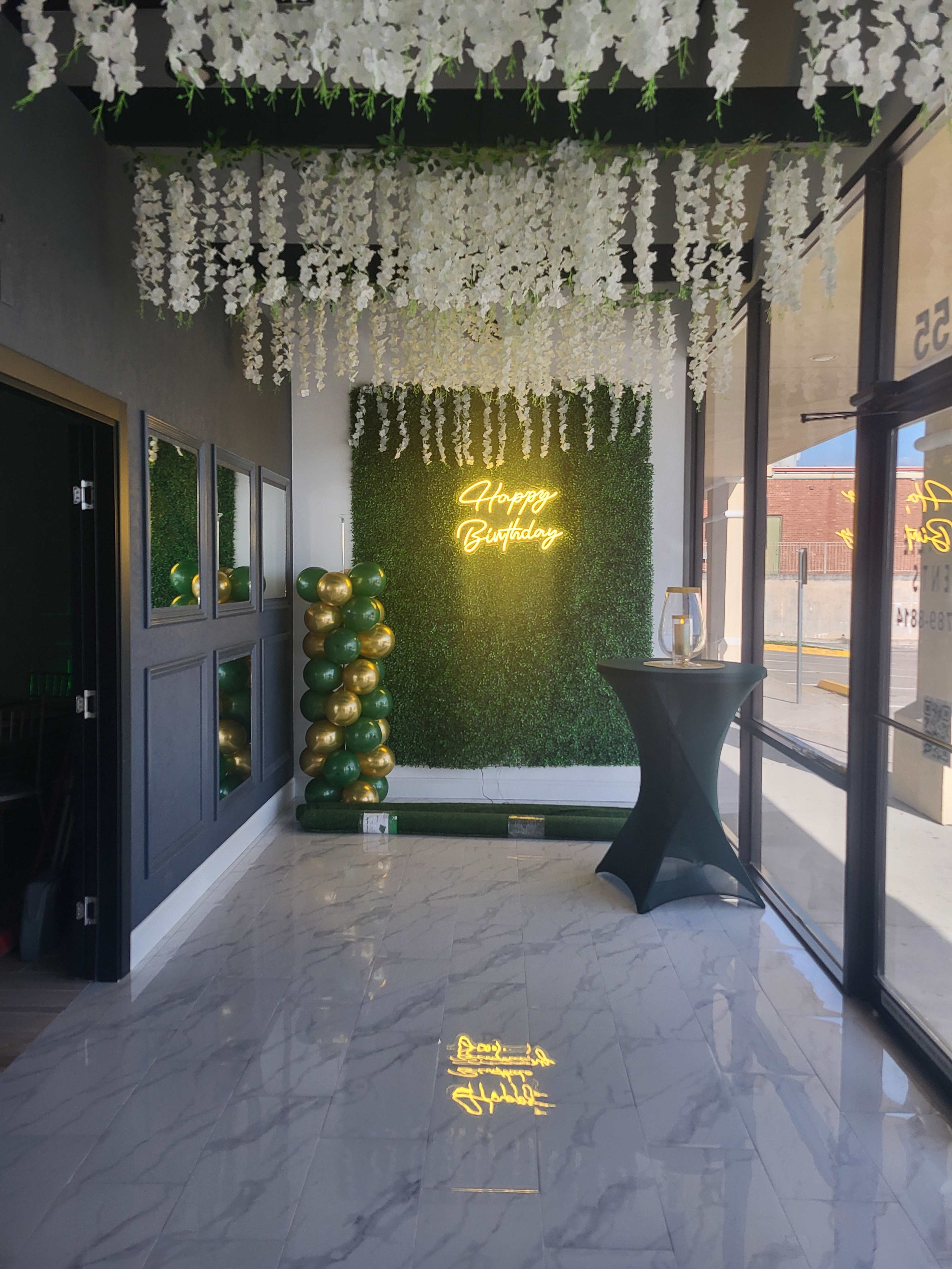 The image shows an event space decorated with greenery and white floral arrangements, featuring a "Happy Birthday" neon sign and a black table in front.