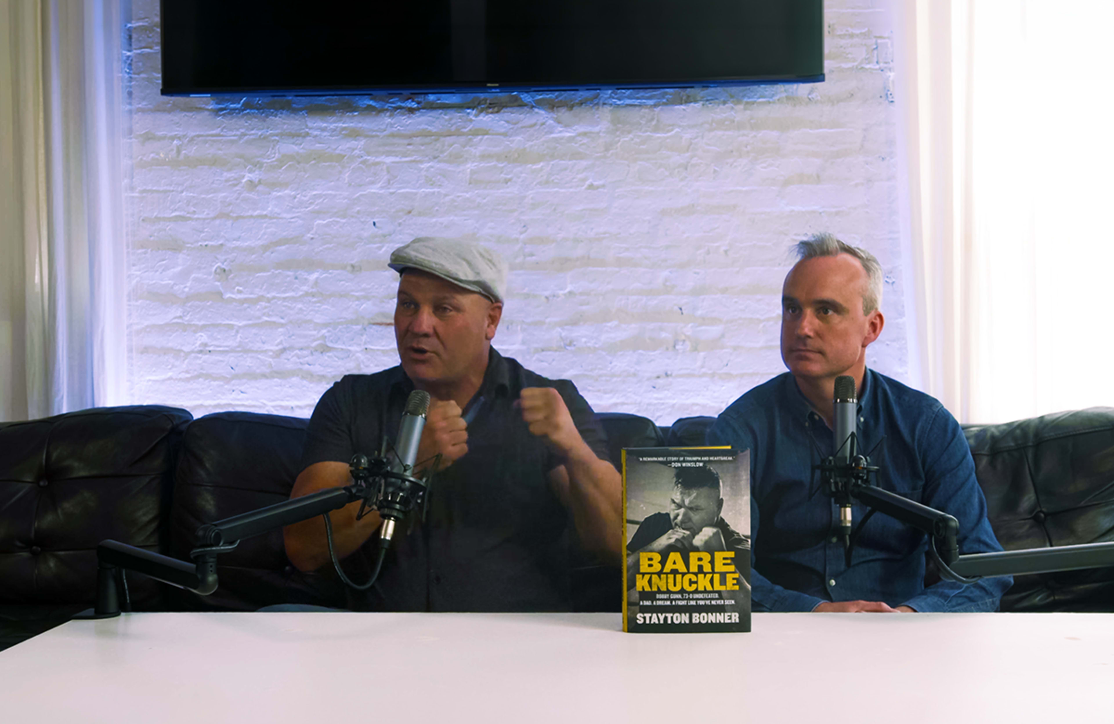 Two men sit at a table with microphones, discussing the book "Bare Knuckle," while a screen displays the title in the background.