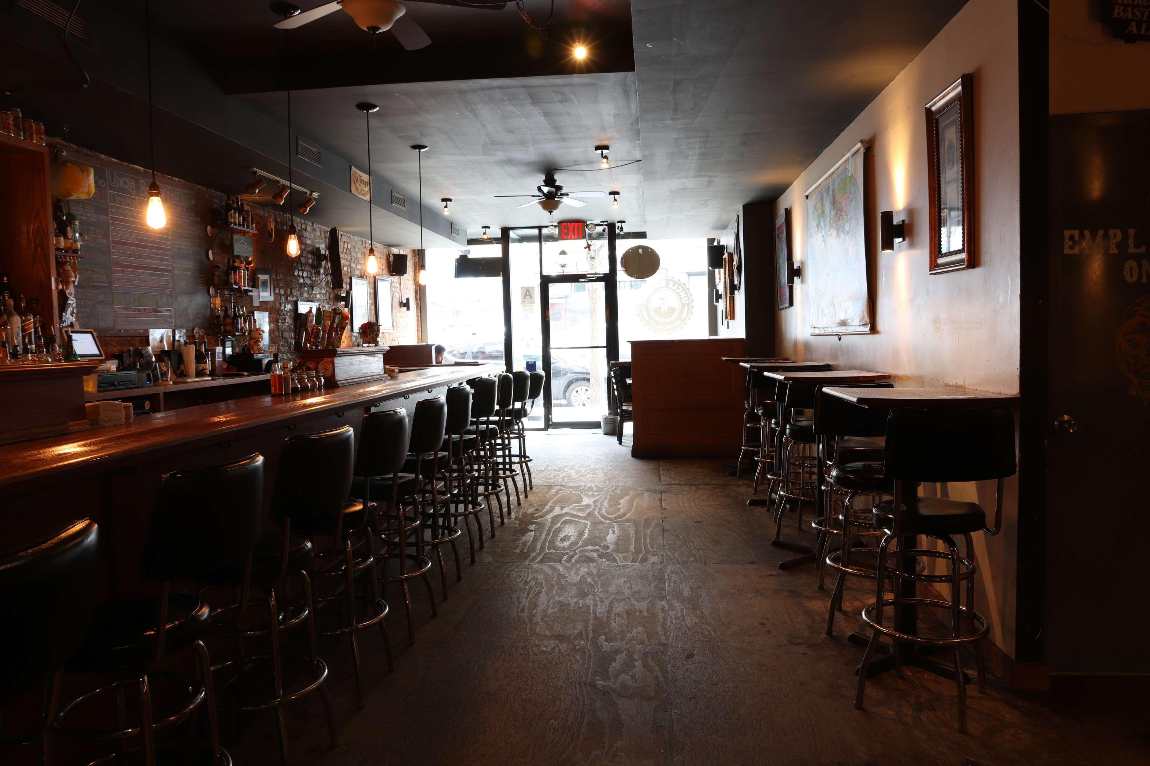 A dimly lit bar with a long wooden counter and high stools, leading to a glass door that opens to the street.