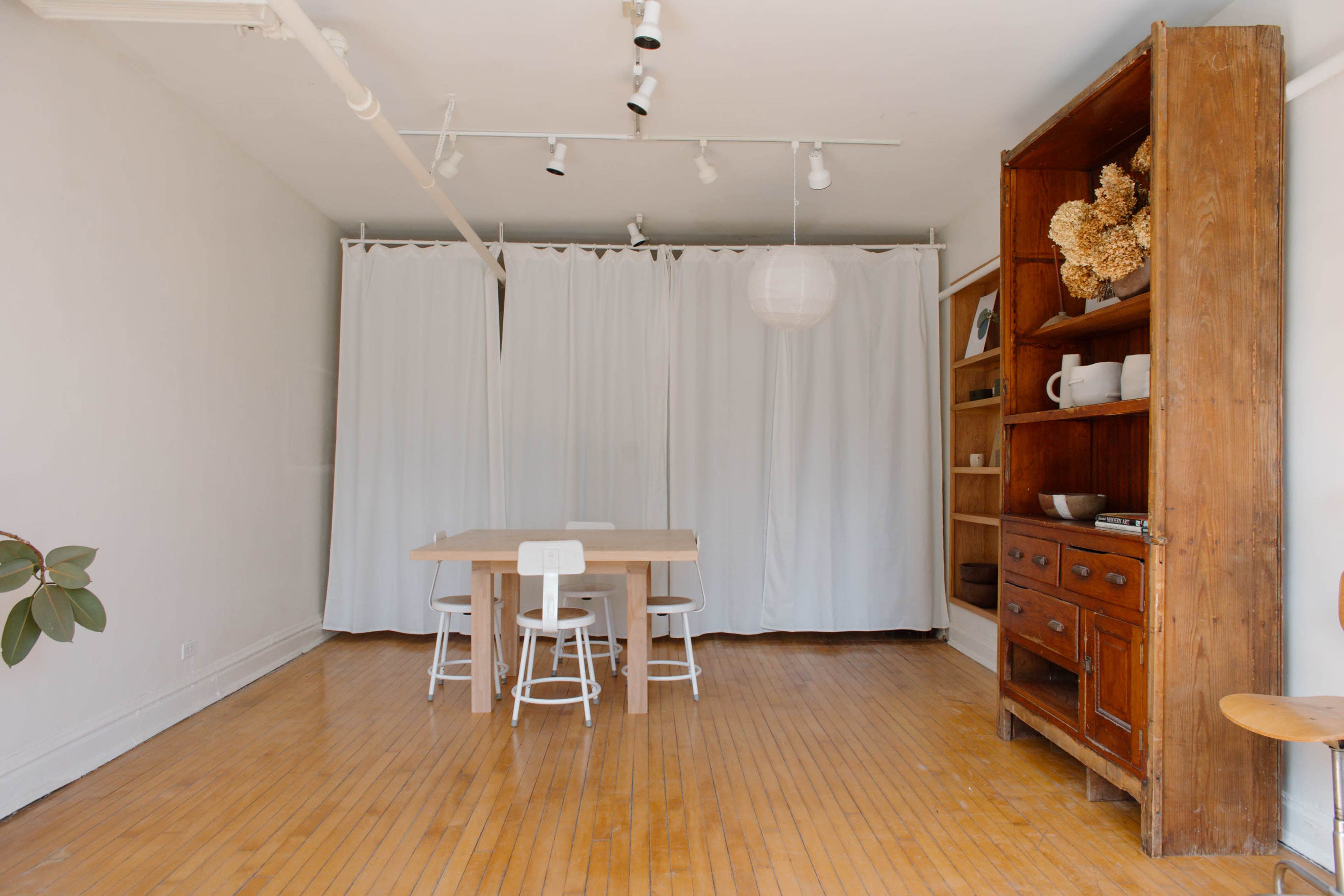 A bright, minimalist room features a wooden dining table with chairs, surrounded by white curtains and a bookshelf against the wall.