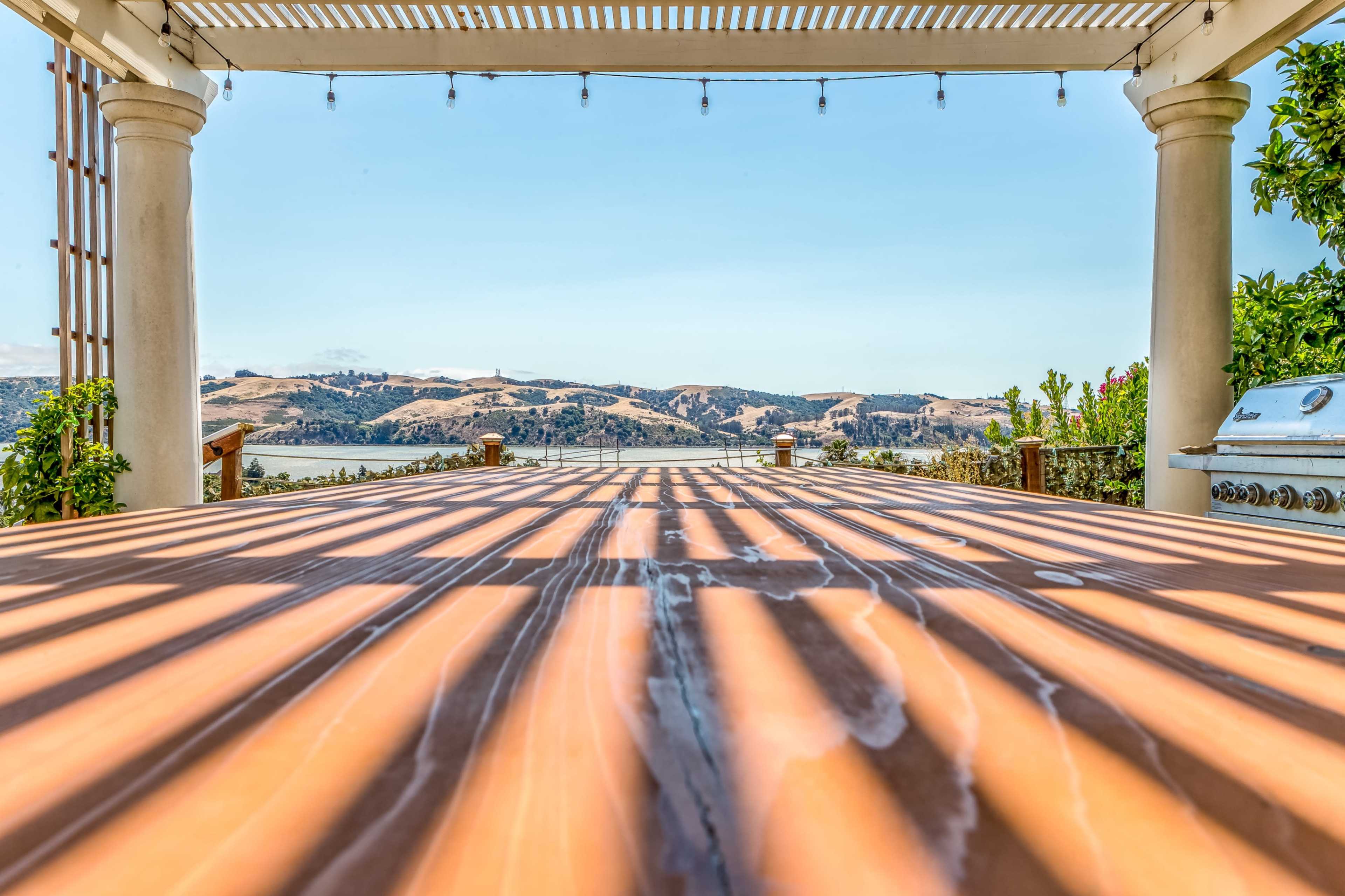 The image shows a wooden table with shadows cast by overhead slats, overlooking a scenic landscape with hills and water in the distance.