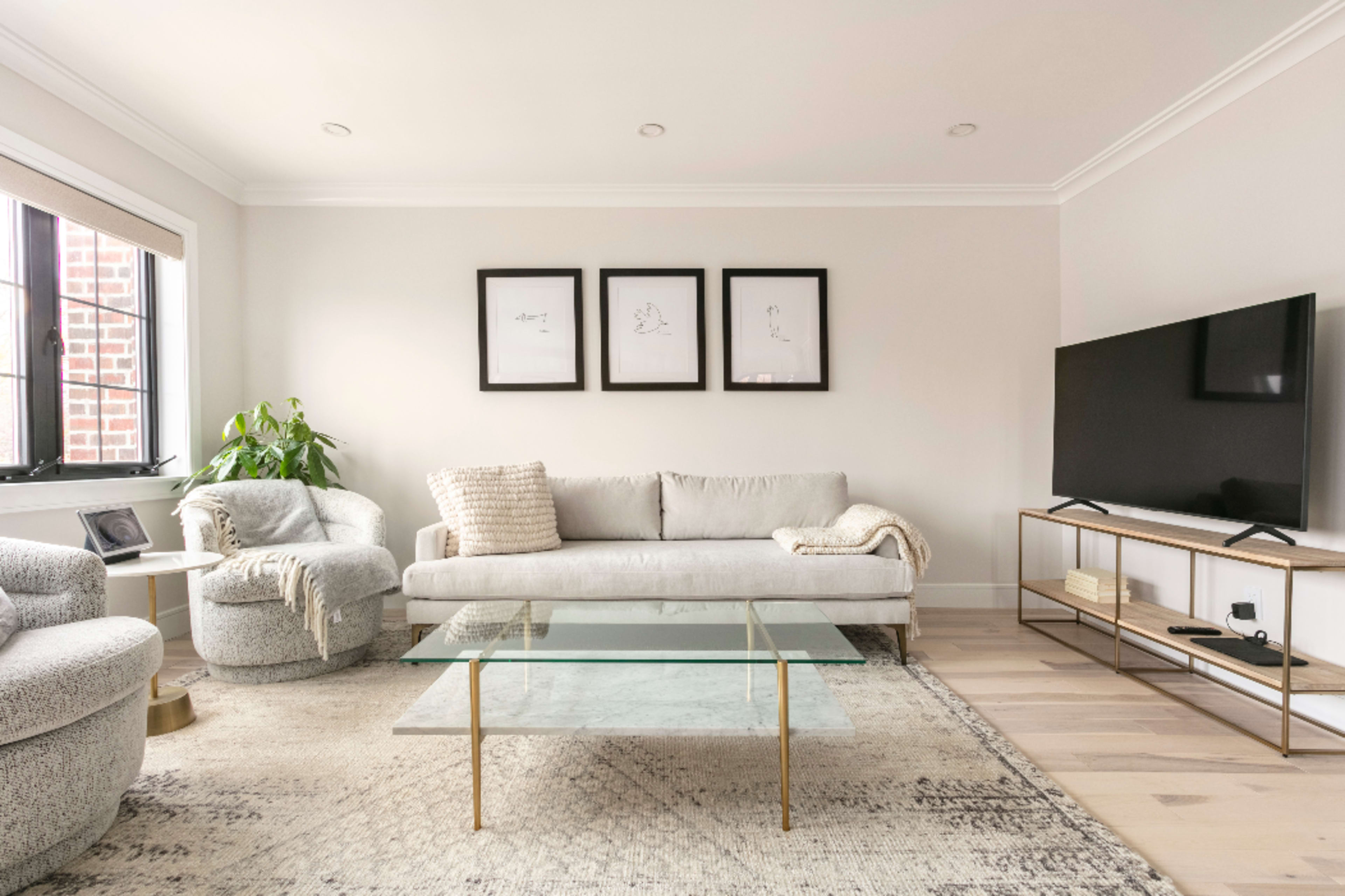 A modern living room features a light-colored sofa, a glass coffee table, a television on a stand, and framed artwork on the walls.