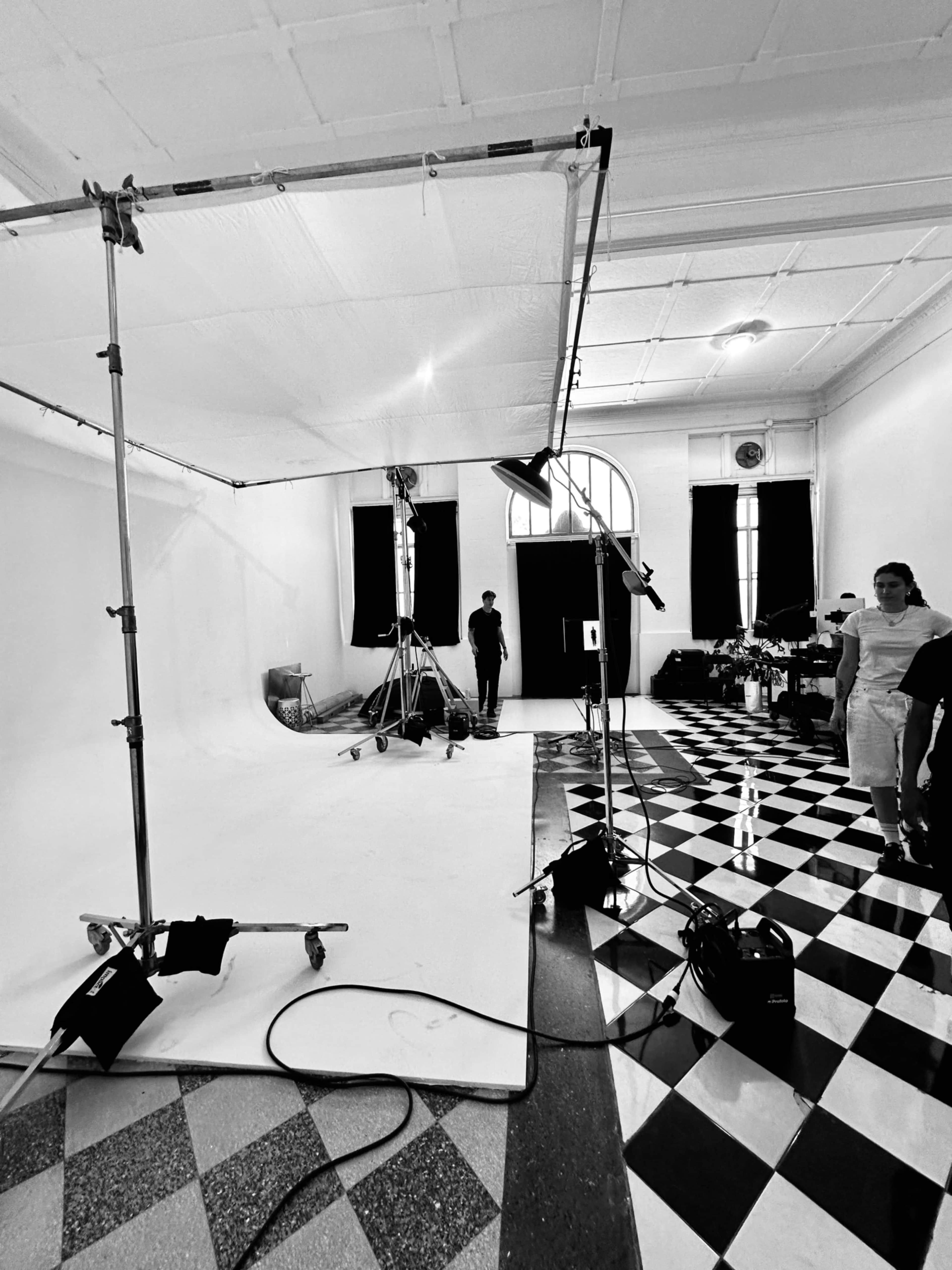 A photography studio features a checkered floor, a large backdrop setup, lighting equipment, and a person standing in the background.