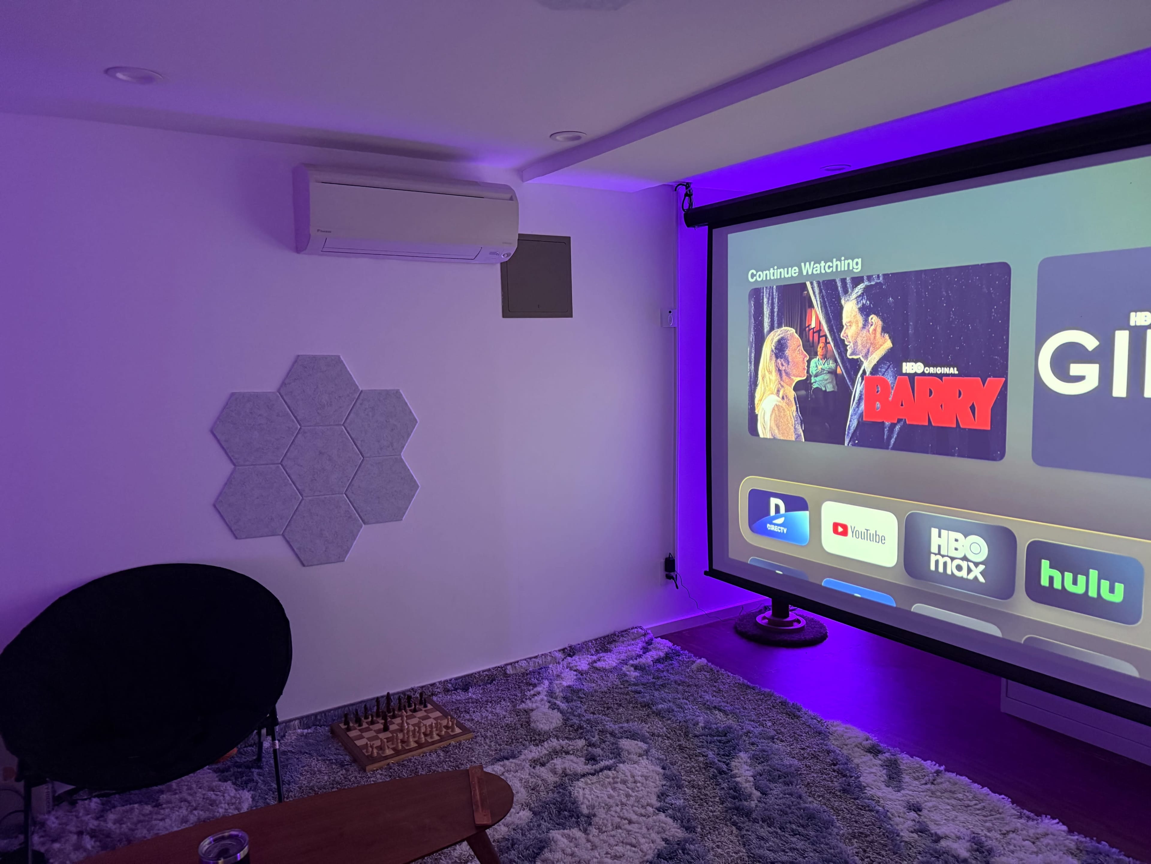 A home theater setup features a large projection screen displaying streaming content, with a black chair, a wooden table, and a chessboard on a patterned rug.