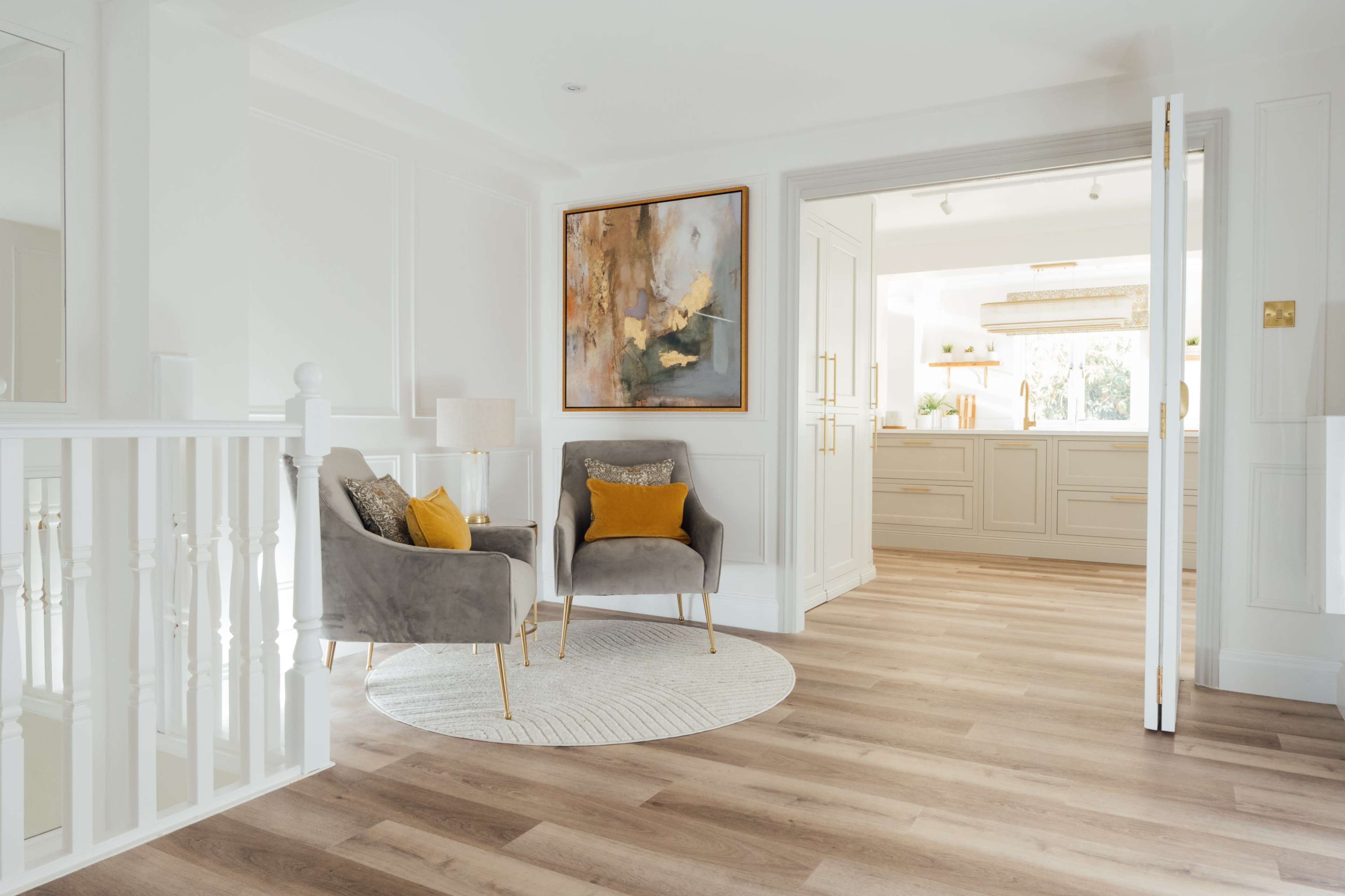 A modern interior with two grey armchairs featuring decorative pillows, a round rug, and an art piece on the wall, leading to a kitchen area.