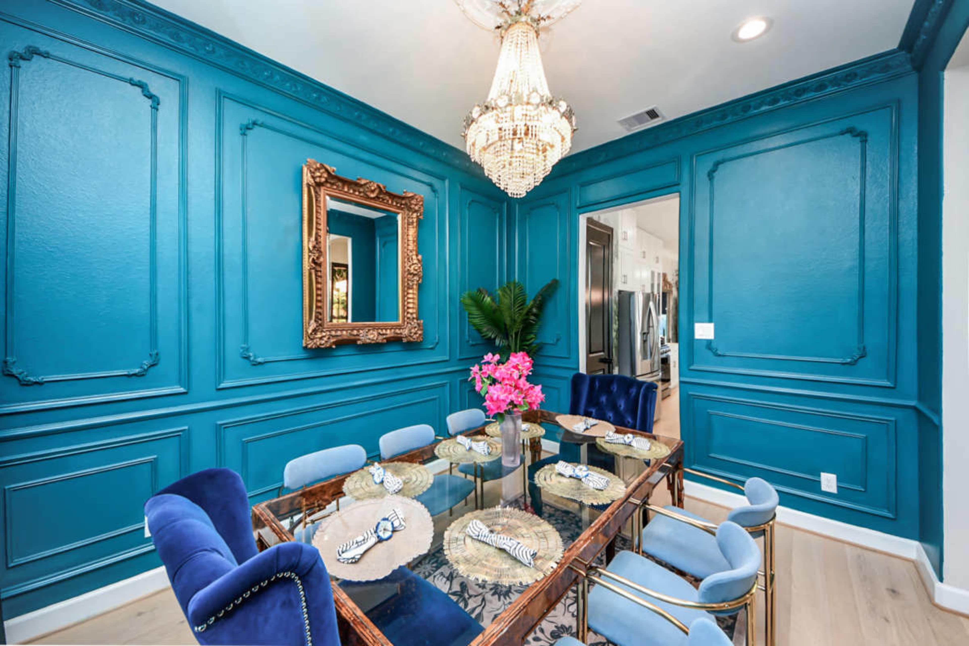 A dining room features a glass table surrounded by blue velvet chairs, with teal walls adorned with decorative molding and a chandelier above.