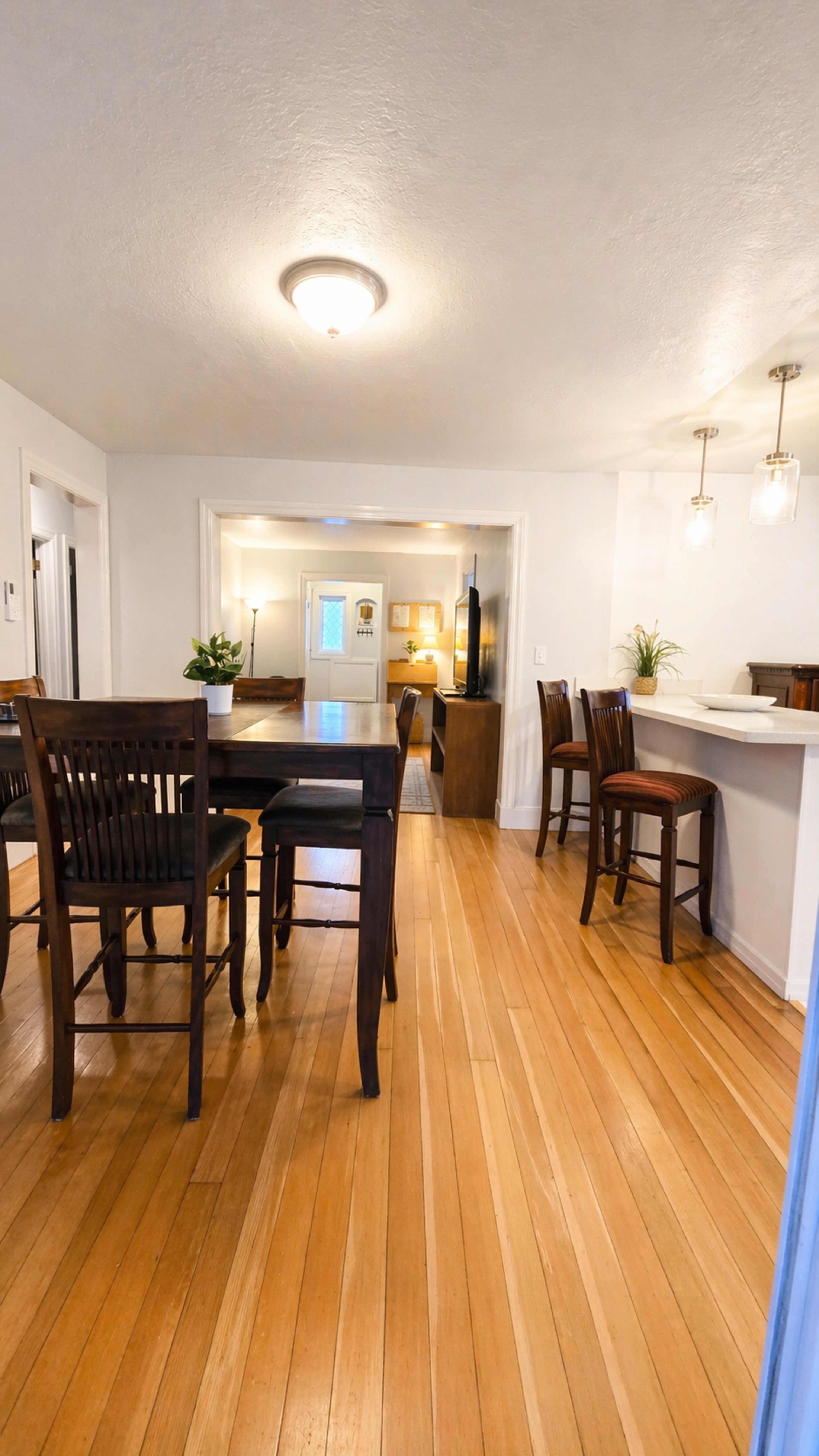 The image shows a bright, open-concept dining and living area with wooden floors and seating arrangements at a table and kitchen counter.