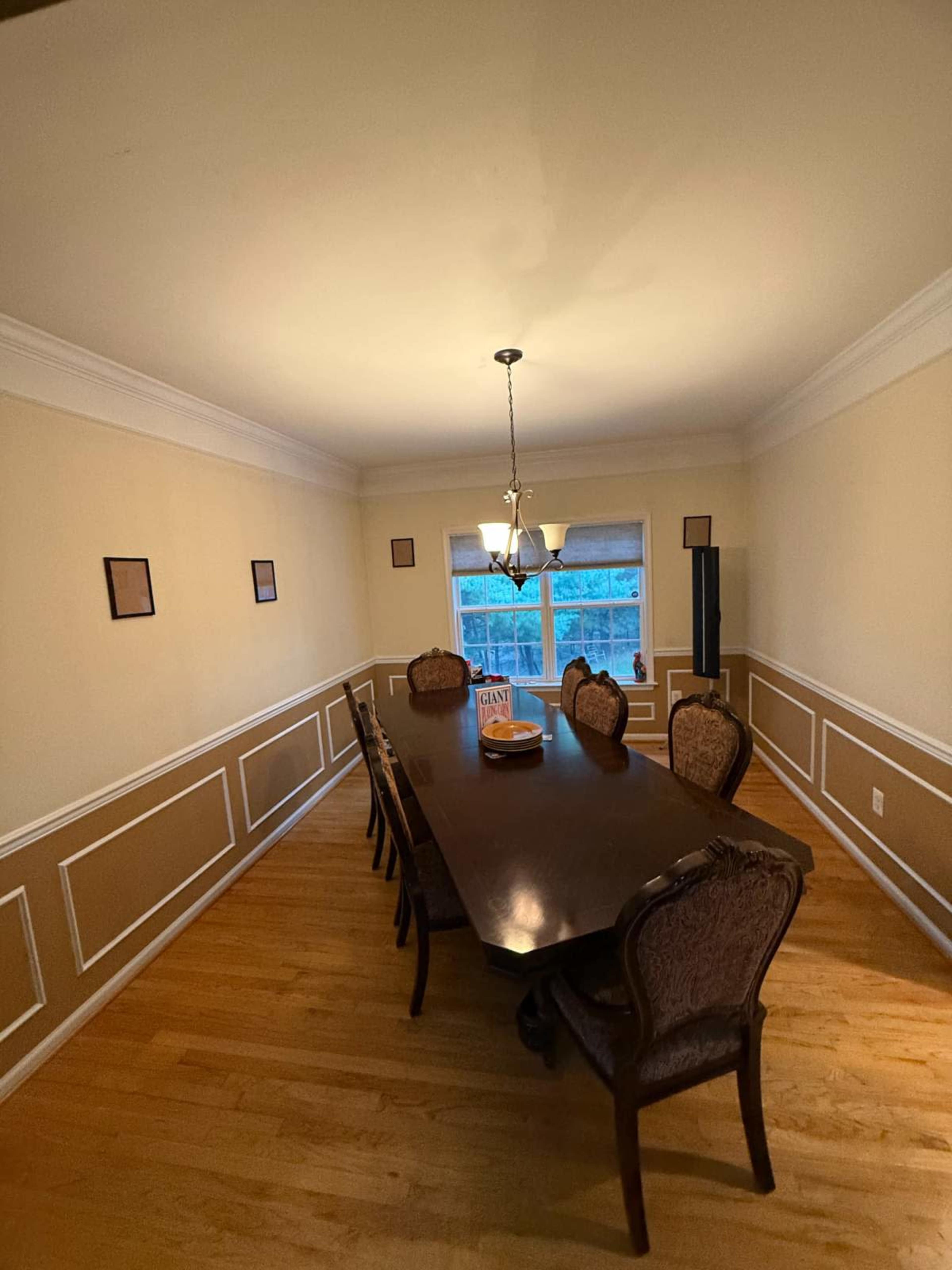 A long dining table with ornate chairs is set in a softly lit room featuring wainscoting and a large window.