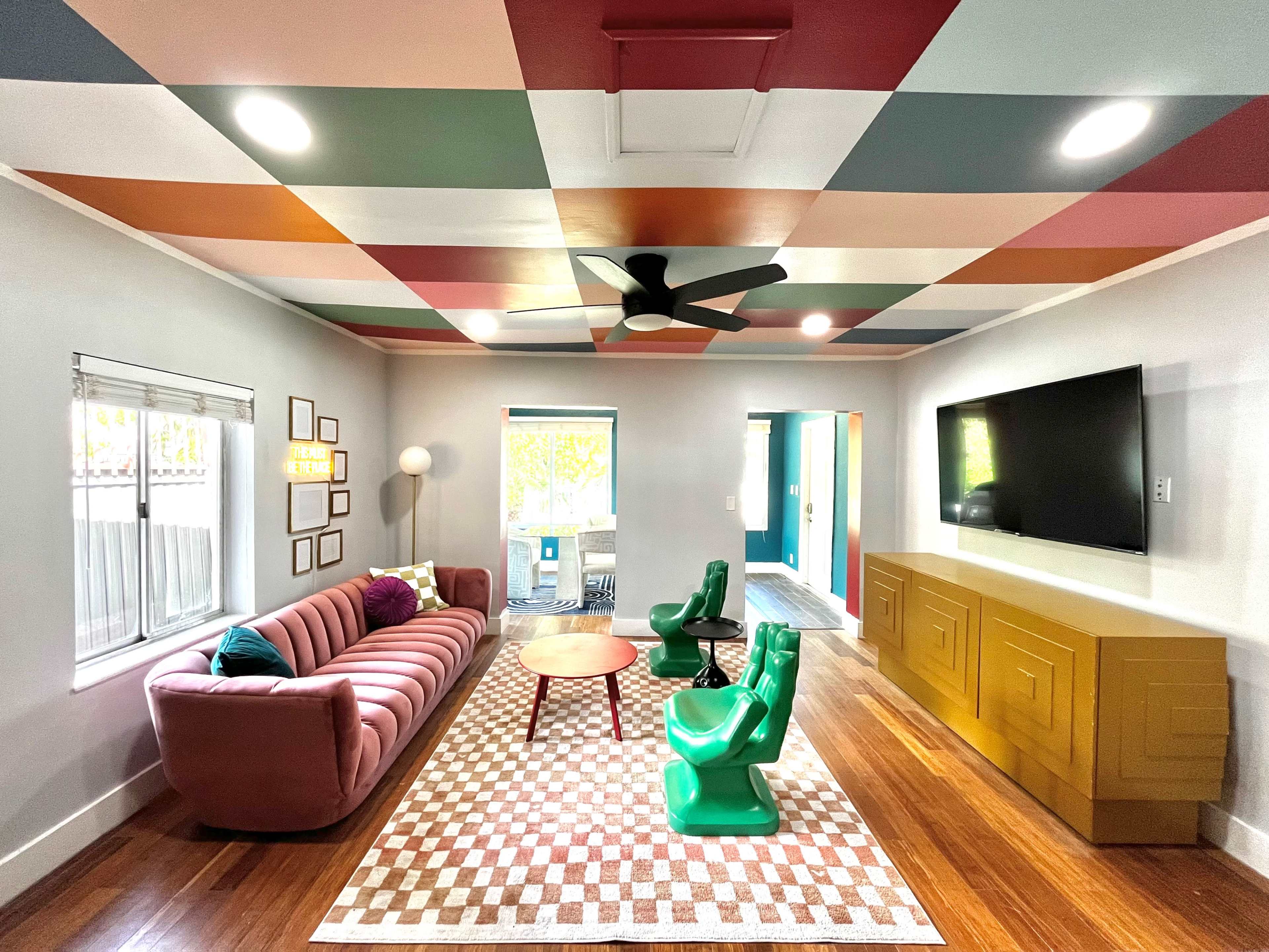 The room features a pink sofa, green chairs, a wooden coffee table, and a colorful ceiling, with large windows allowing natural light to enter.