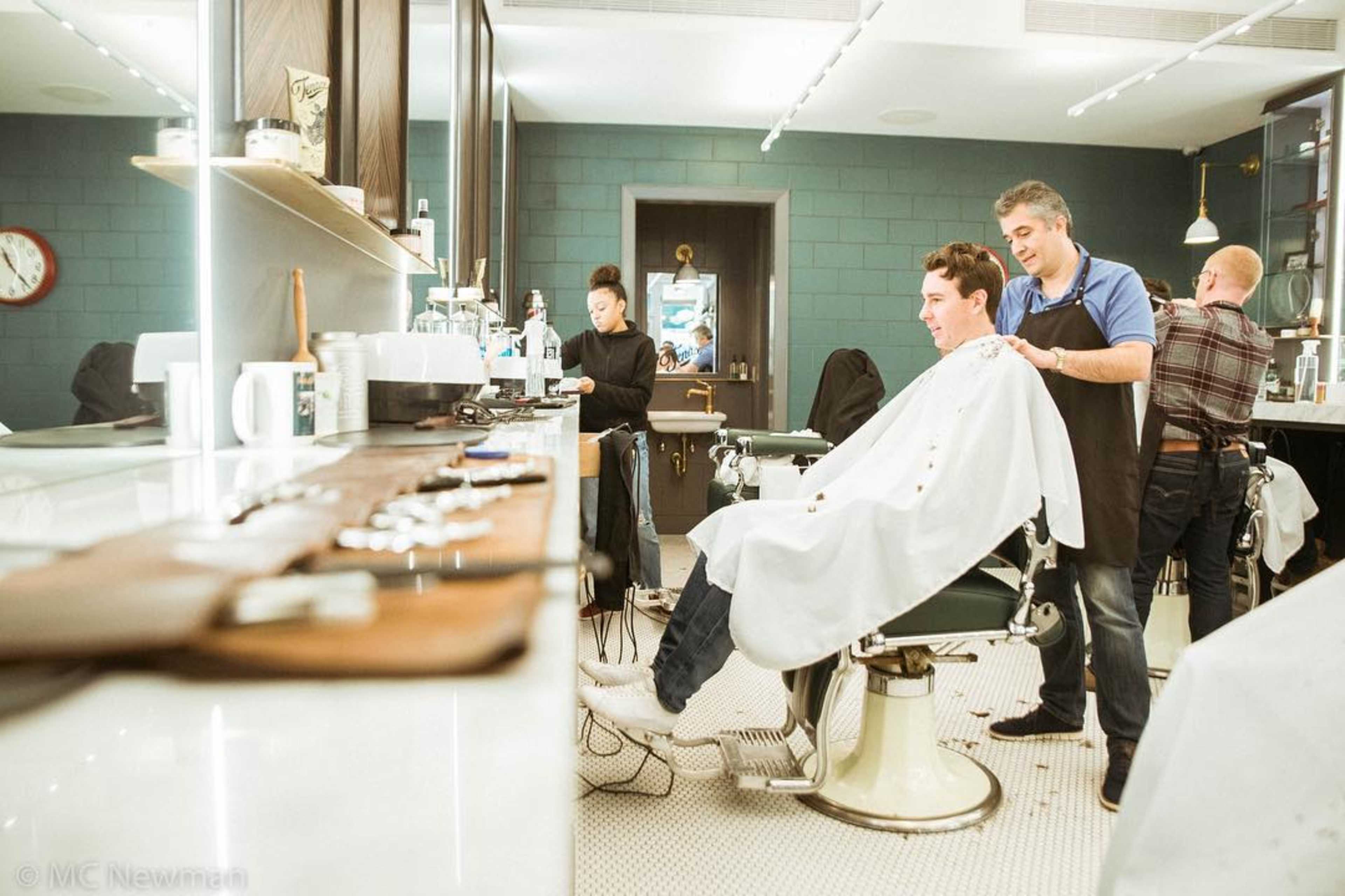 A barber styles a client's hair while another stylist attends to a different customer in a modern barbershop.