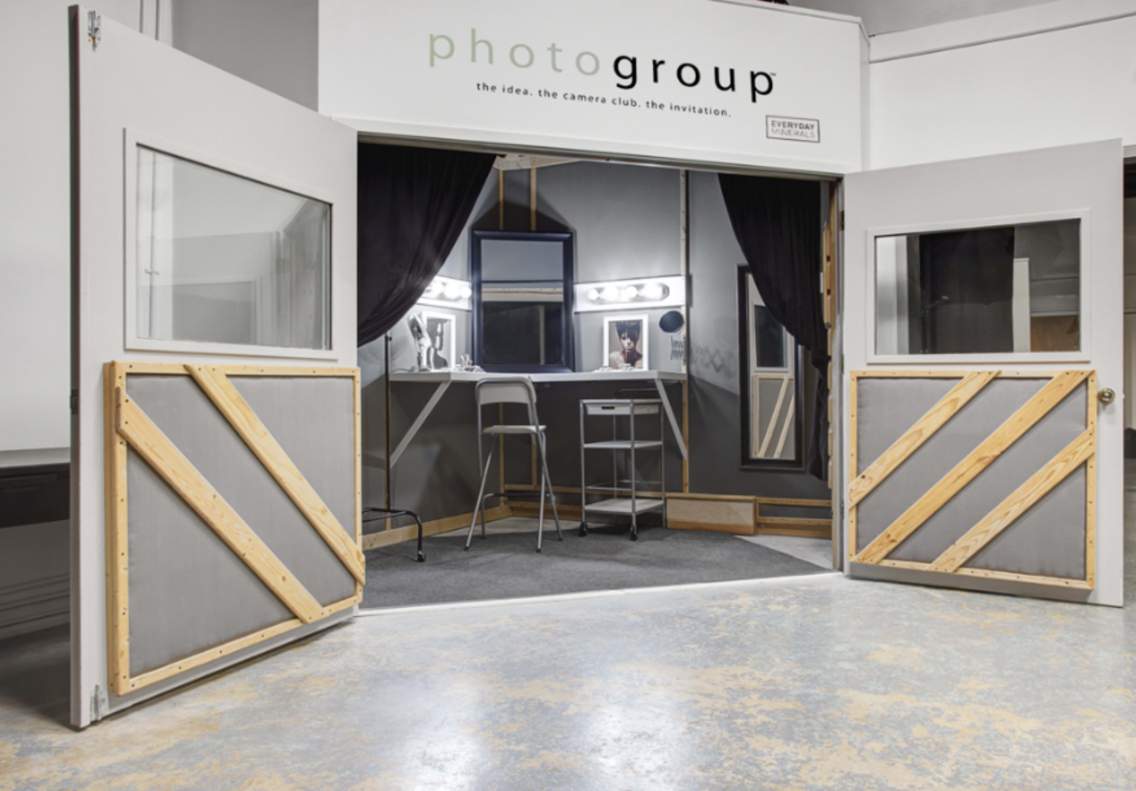 An entrance to a studio space called "photogroup," featuring double doors with wooden cross bracing and a makeup area visible inside, illuminated by vanity lights.