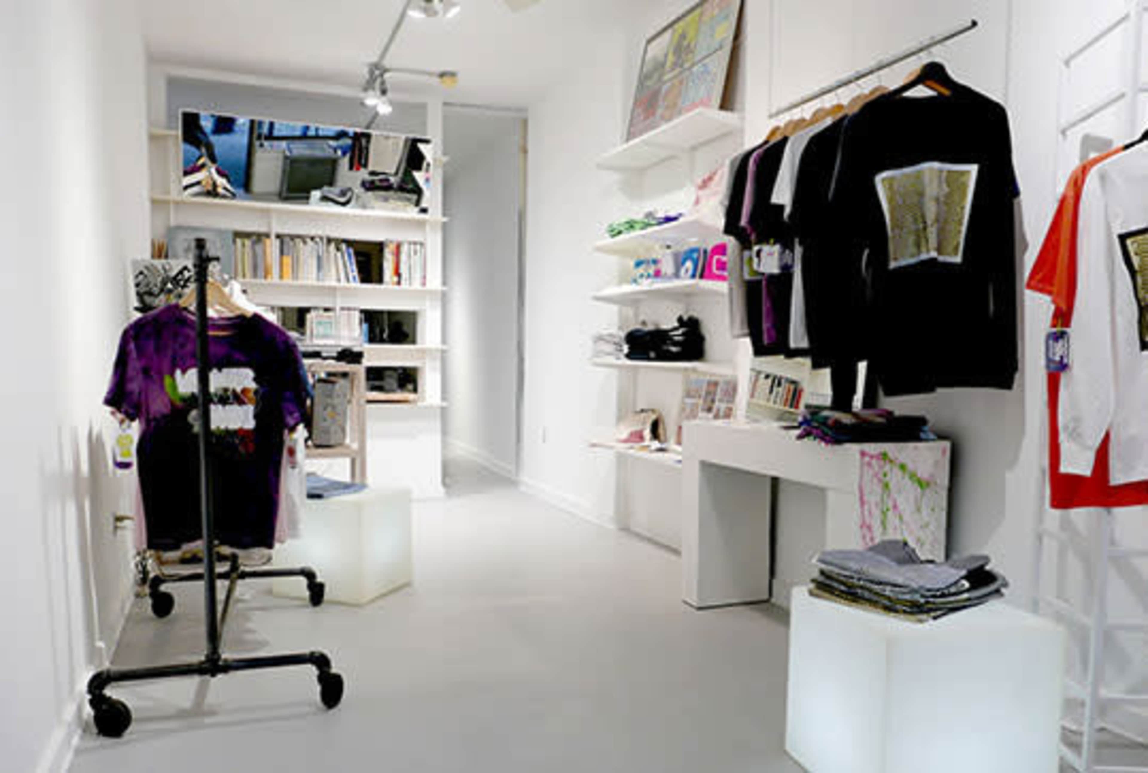 A clothing boutique with a minimalist white interior, featuring racks of t-shirts, shelves of books, and display tables.