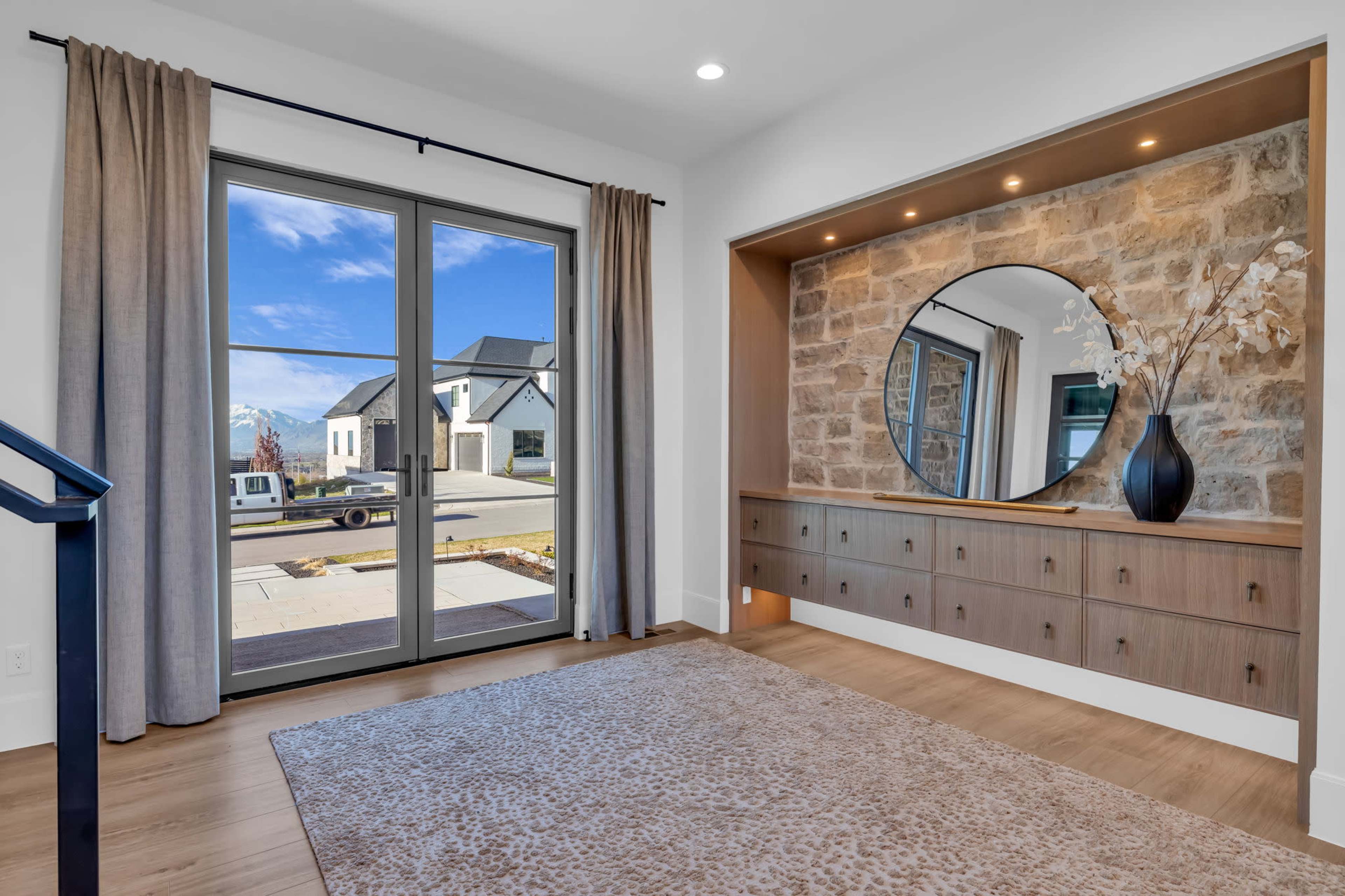 The image shows a modern entryway with a large round mirror, a wooden console with drawers, a rug on the floor, and large double doors leading to a view of the exterior landscape.