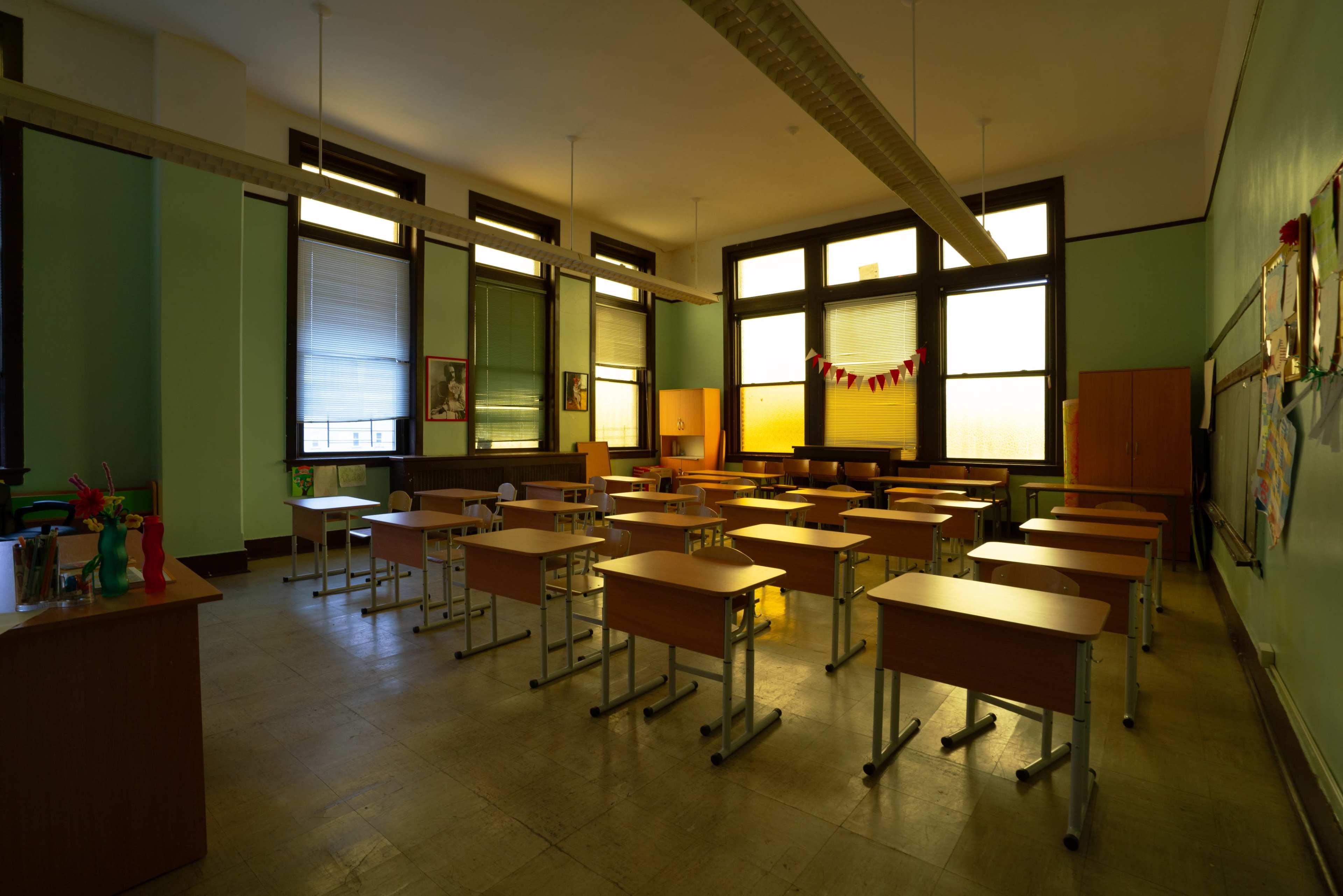 A vacant classroom features rows of wooden desks and large windows, with green walls and minimal decorations.