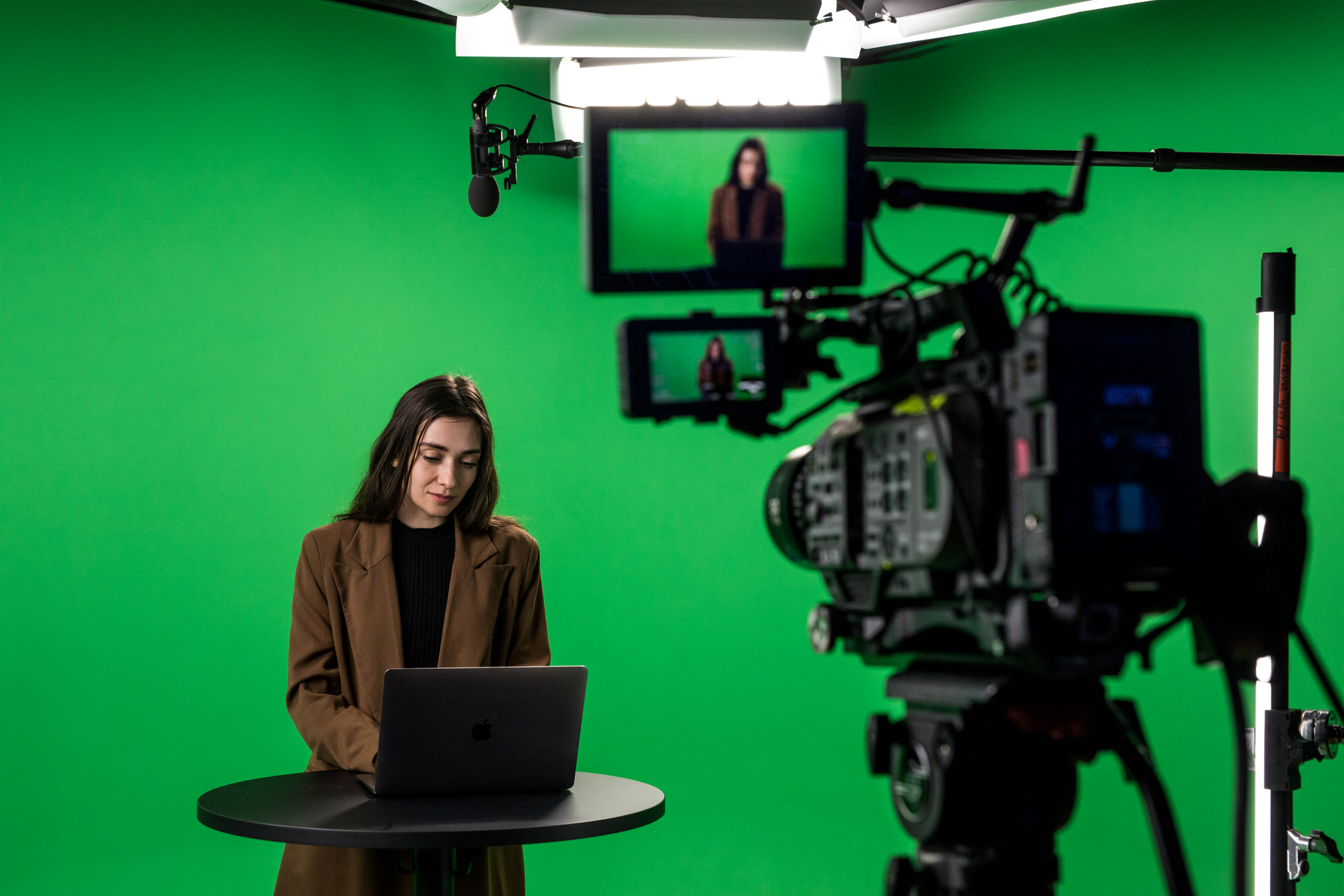 A woman is sitting at a table with a laptop in front of her, as a camera and monitors capture her in a studio with a green backdrop.