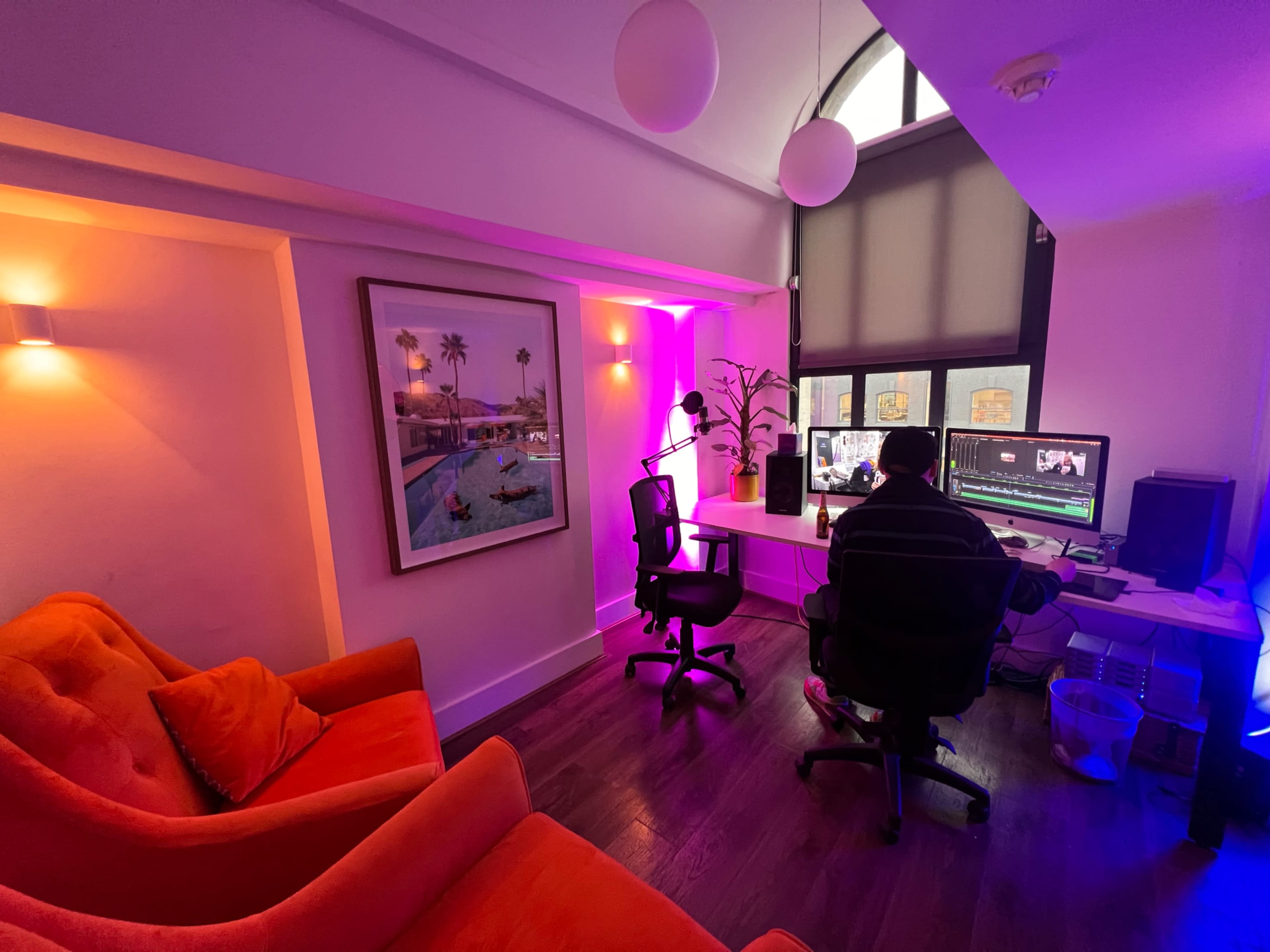 The image shows a brightly lit workspace with a person editing video on a computer, surrounded by orange seating and colorful ambient lighting.
