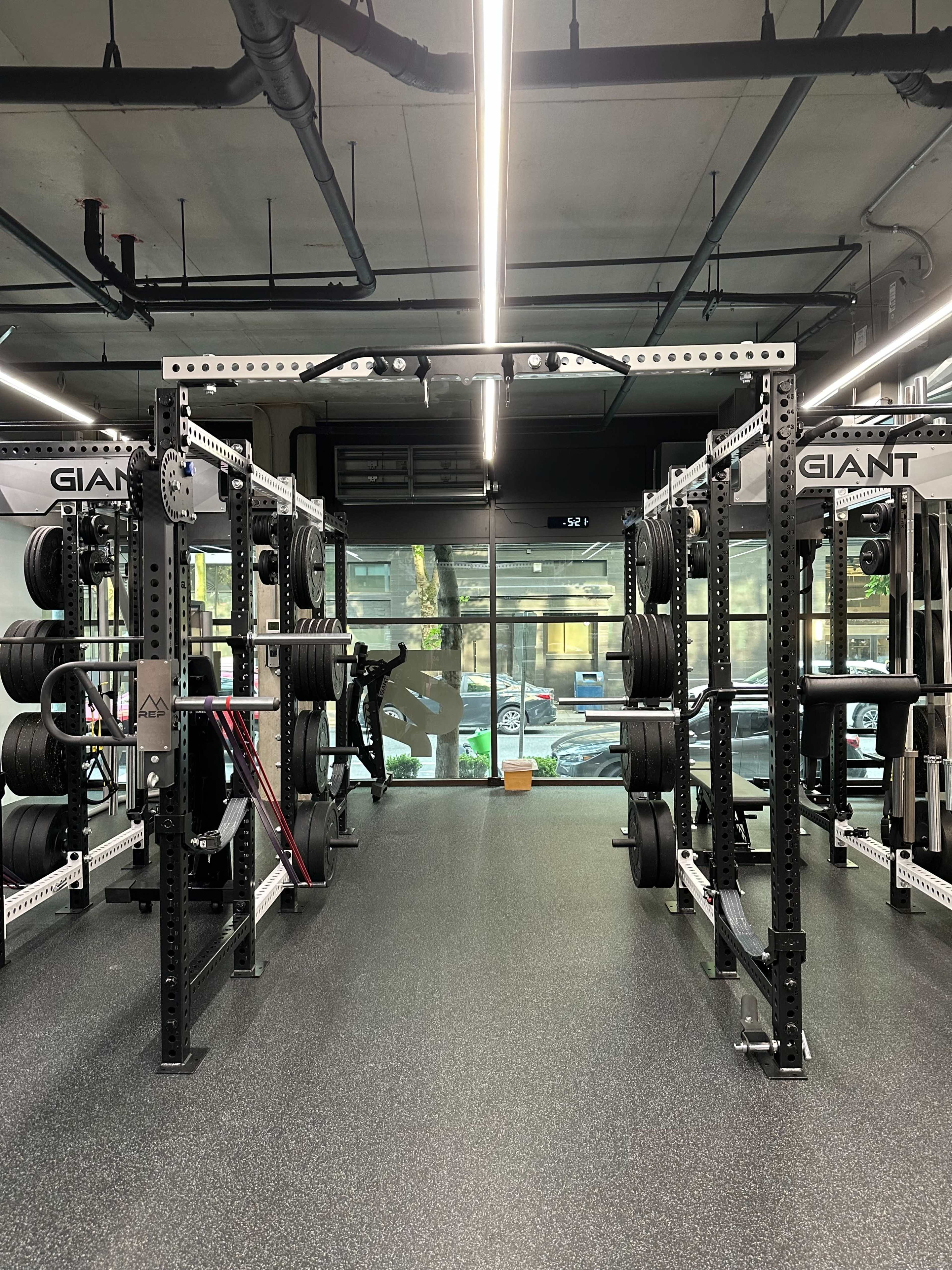 The image shows a modern gym interior with weightlifting racks and large windows that overlook an outside street.