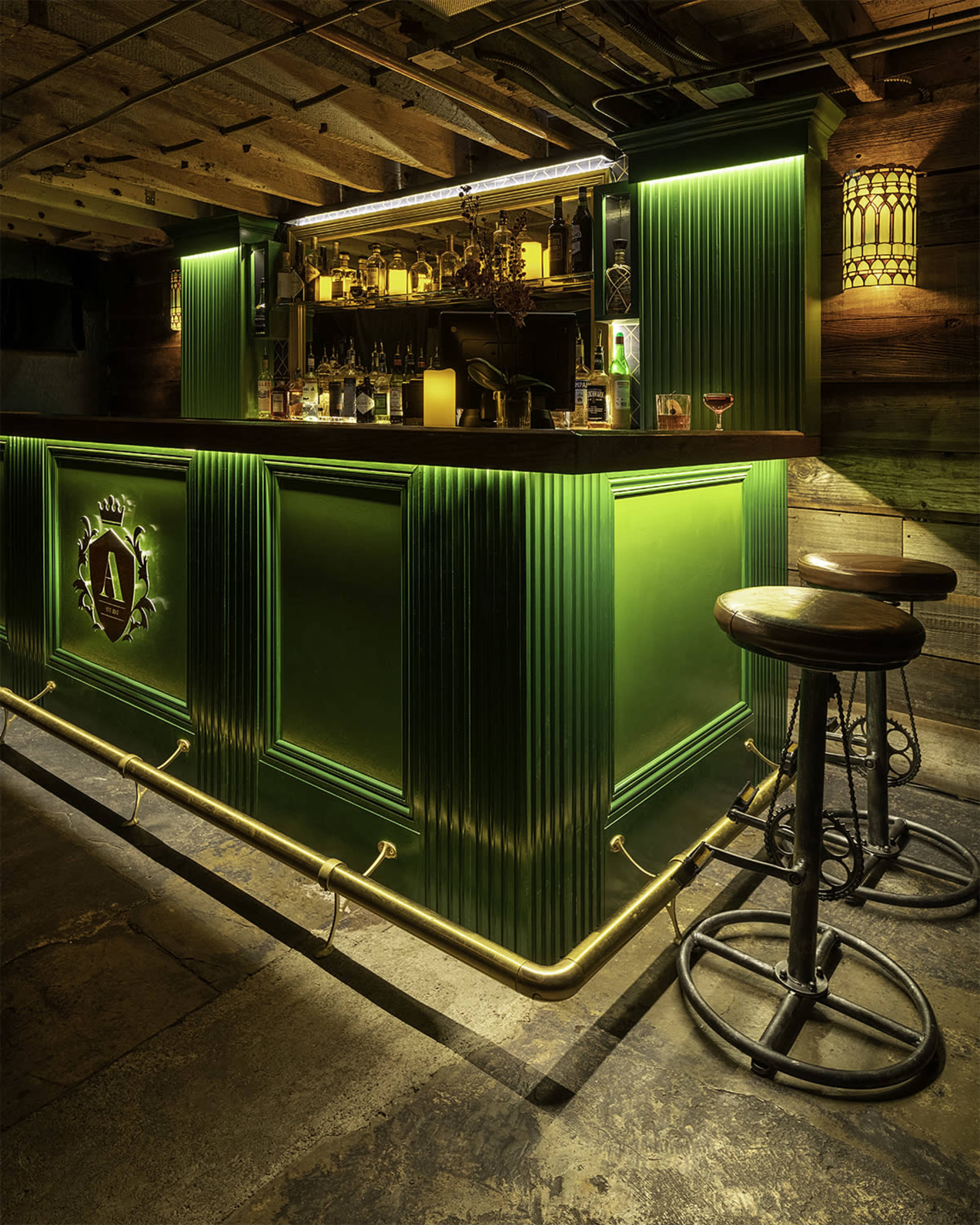 A green bar with a well-stocked shelf of liquor bottles illuminated by overhead lighting in a rustic setting.