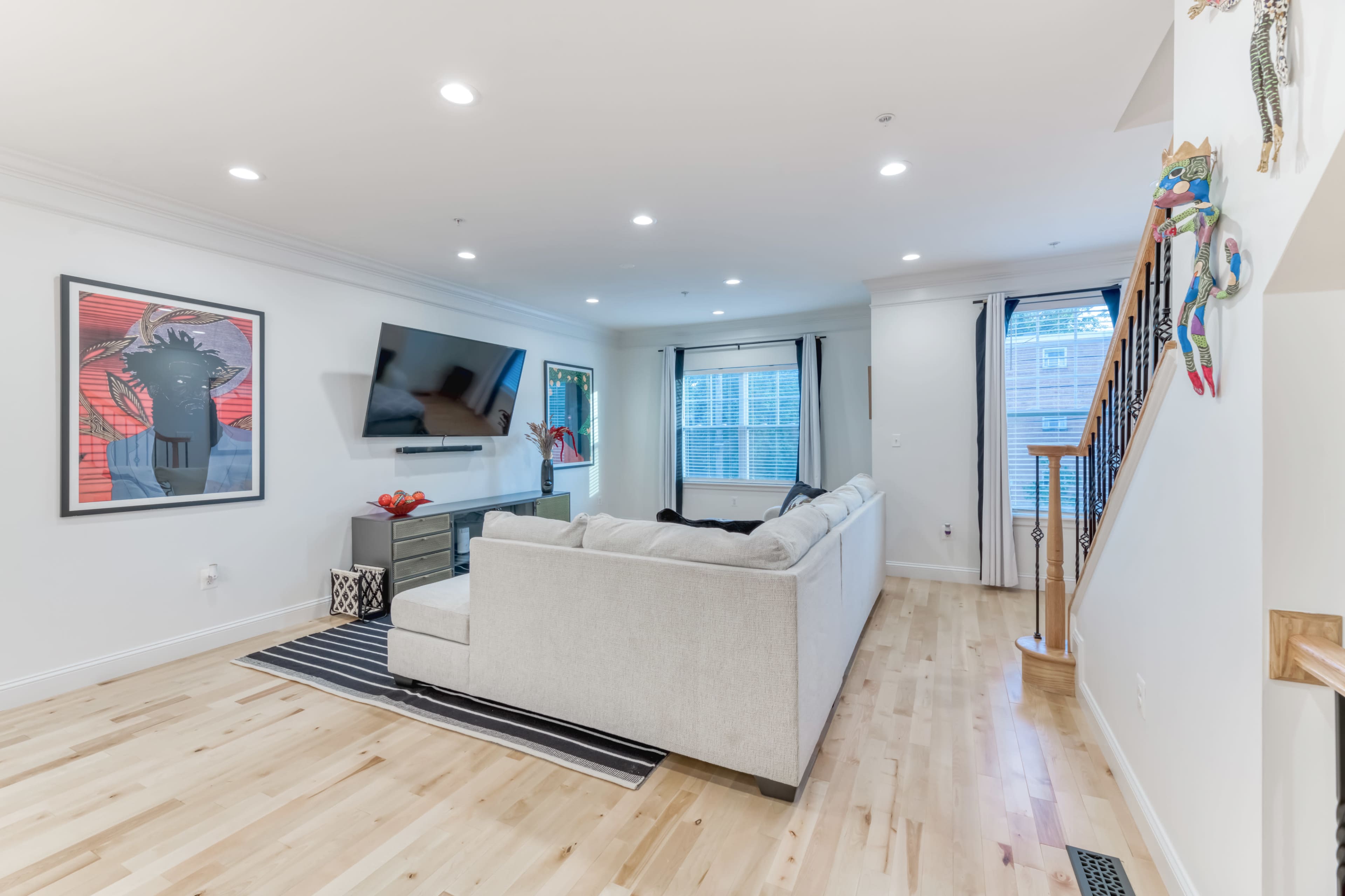 The living room features a light-colored sectional sofa, a mounted television on the wall, and a wooden staircase in the background.