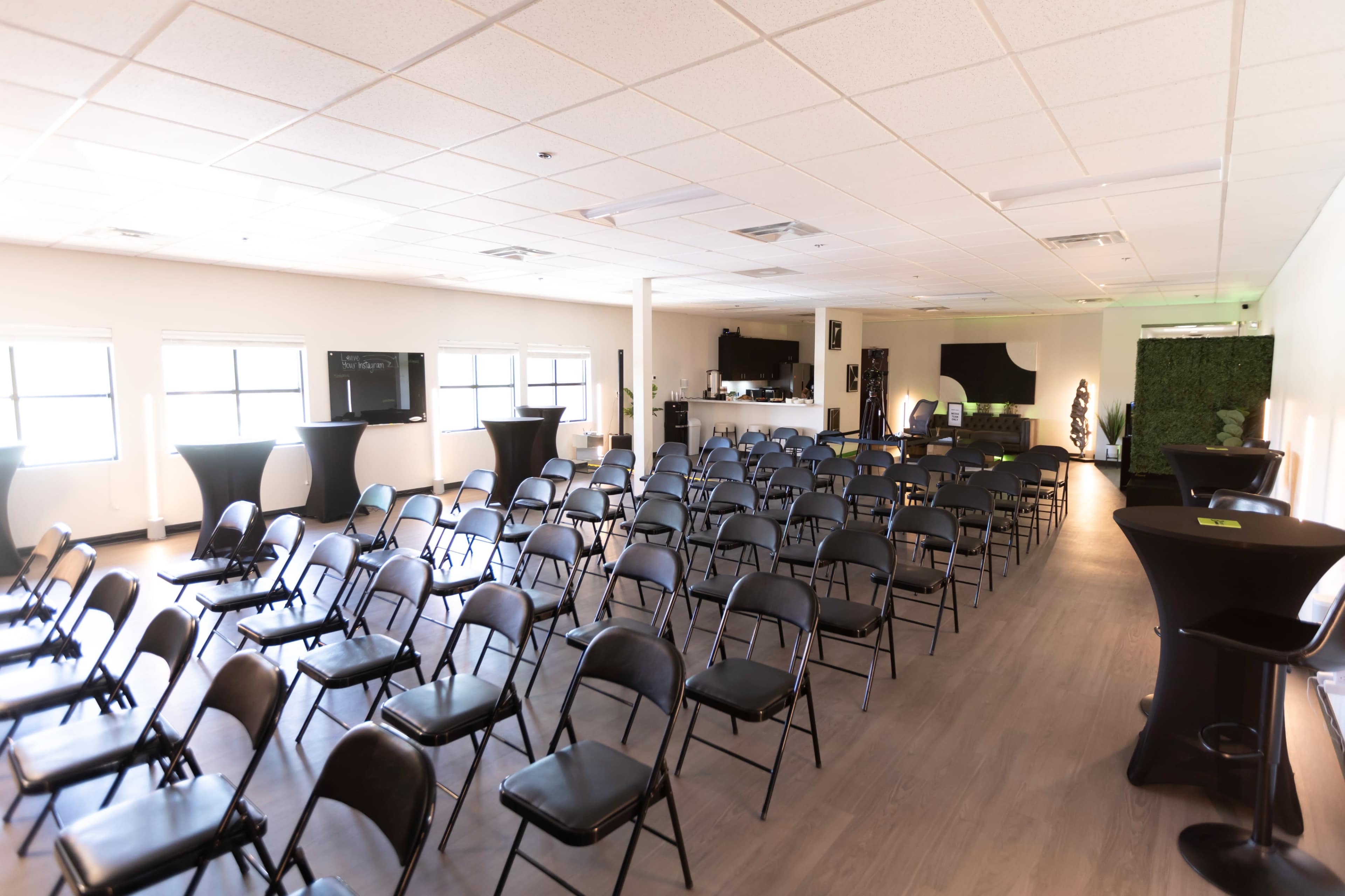 A spacious room arranged with rows of black chairs facing a service area and several tall tables set up around the perimeter.