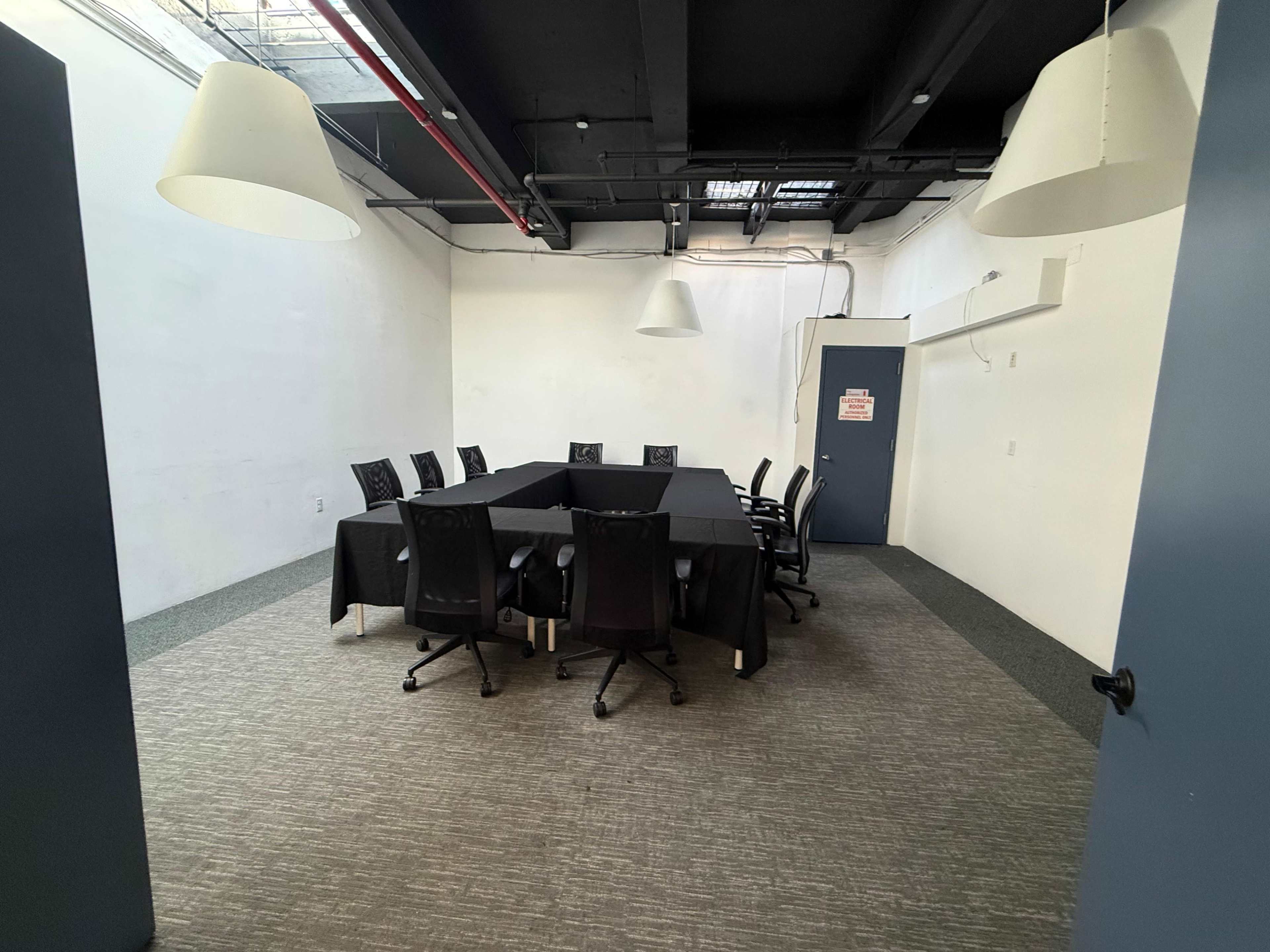 A conference room features a long rectangular table surrounded by black chairs, with a grey carpeted floor and two overhead pendant lights.