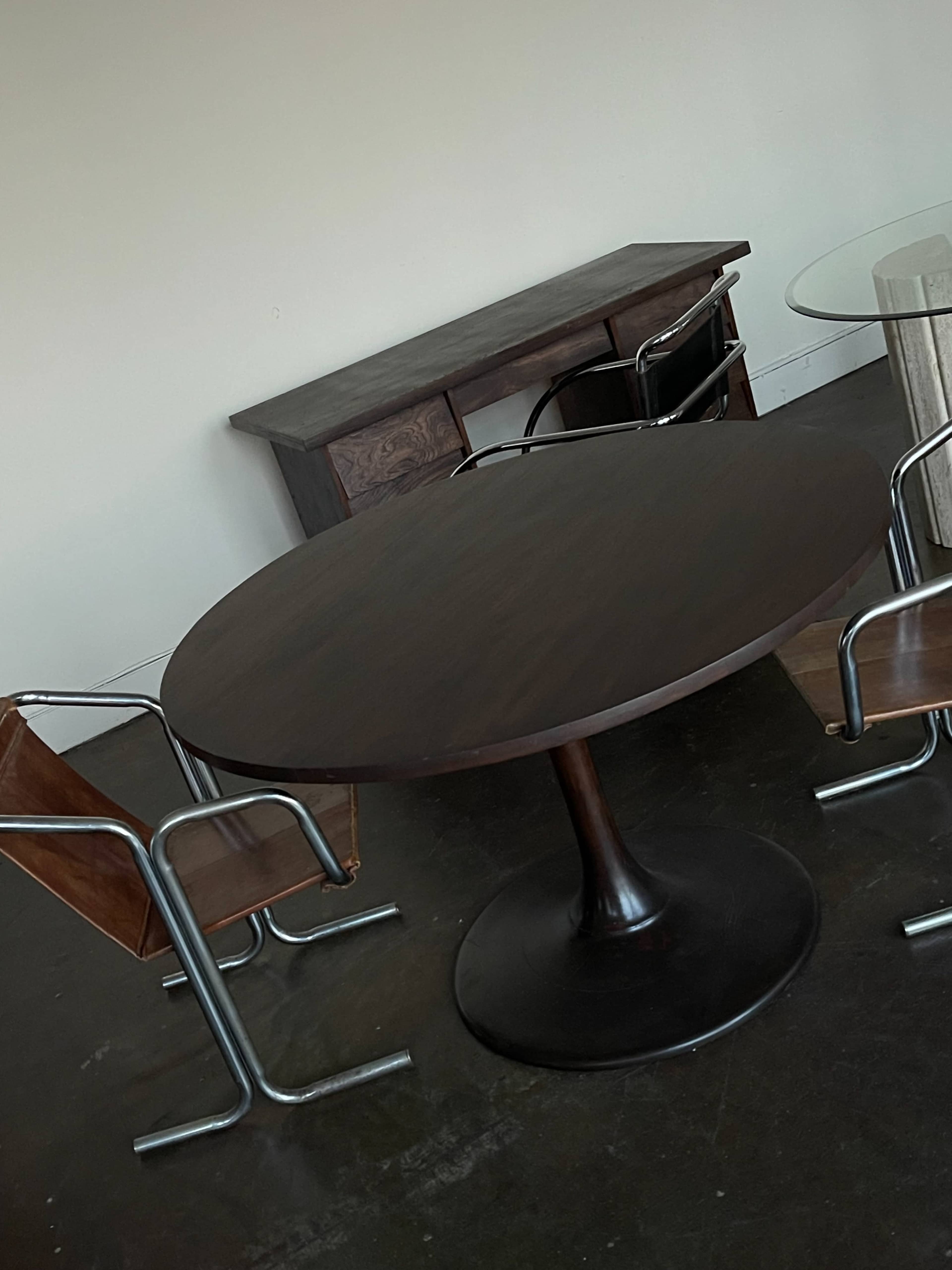 The image shows a minimalist room with a round, dark wooden table surrounded by four modern chairs, including two with metal frames and wooden seats.