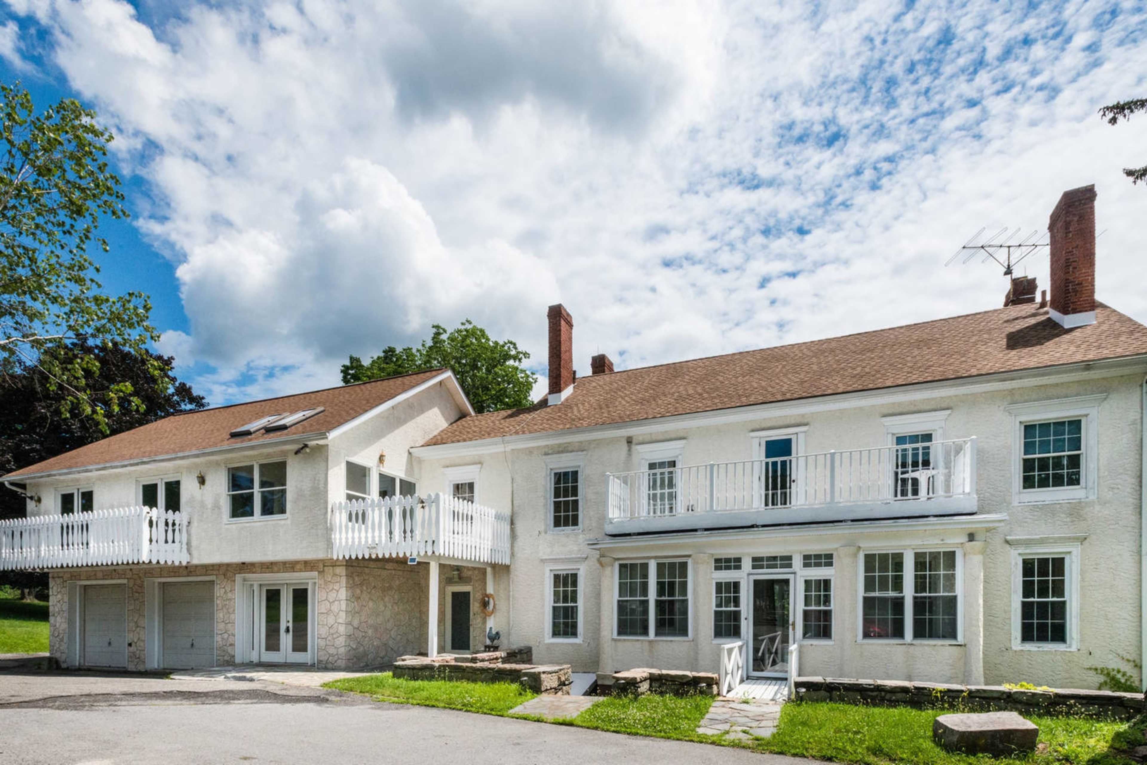 The image shows a large, two-story house with a raised balcony and a separate, attached garage on one side.