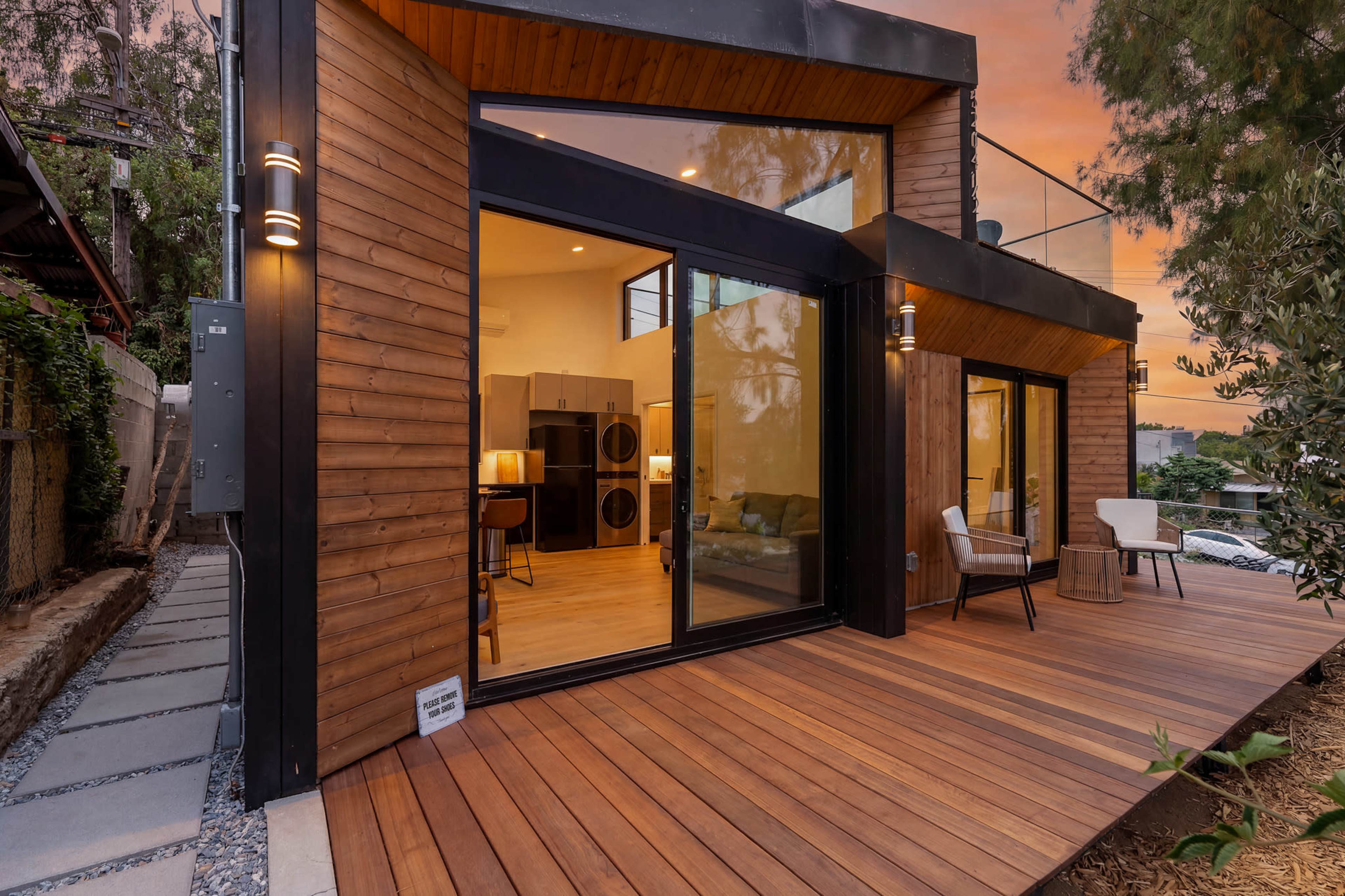 The image shows a modern wooden house with large glass sliding doors opening onto a wooden deck, surrounded by greenery and rocky landscaping.