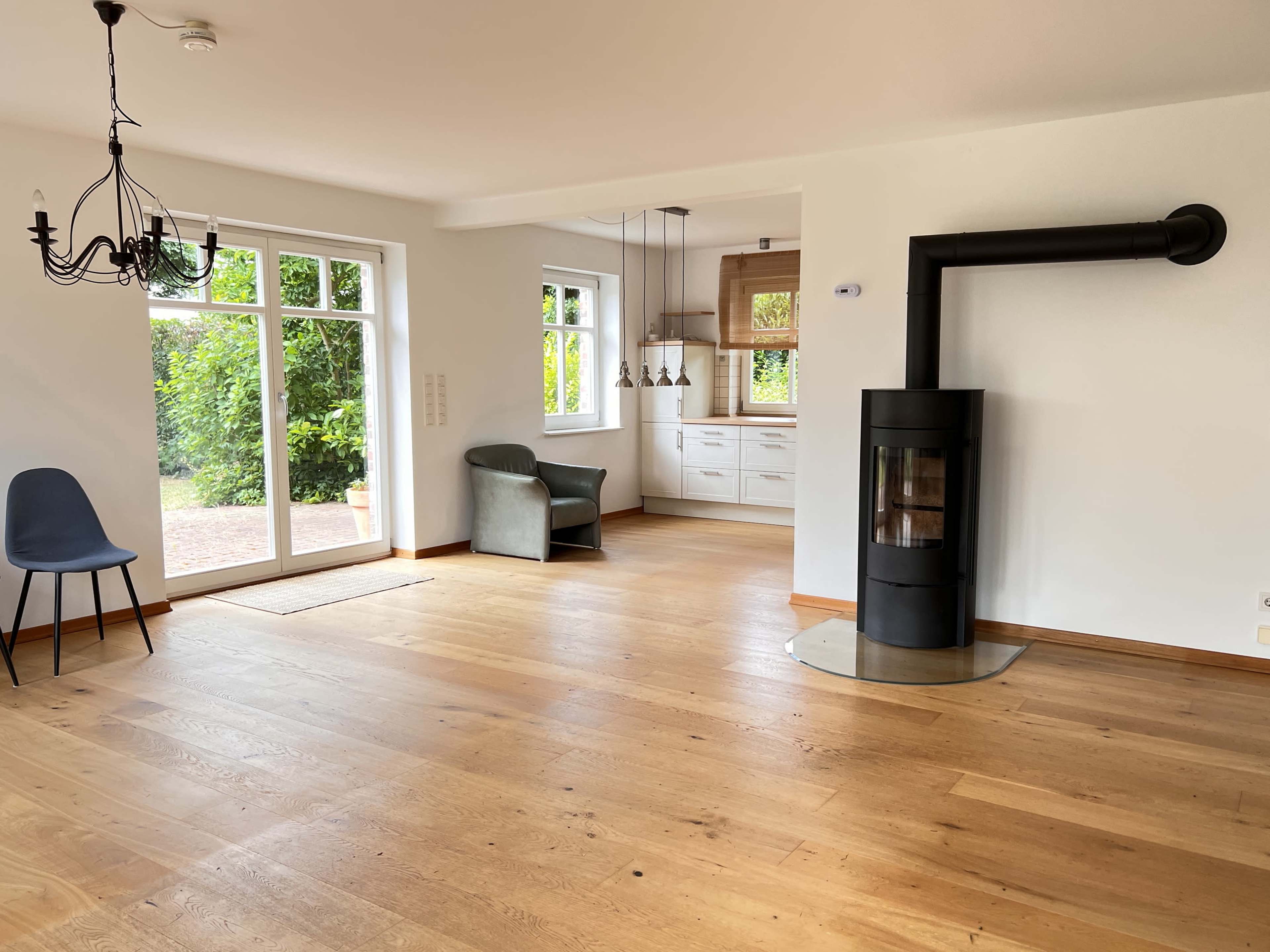 The image shows a spacious, bright interior featuring a living area with wooden flooring, a modern black stove, and a kitchen area visible in the background.