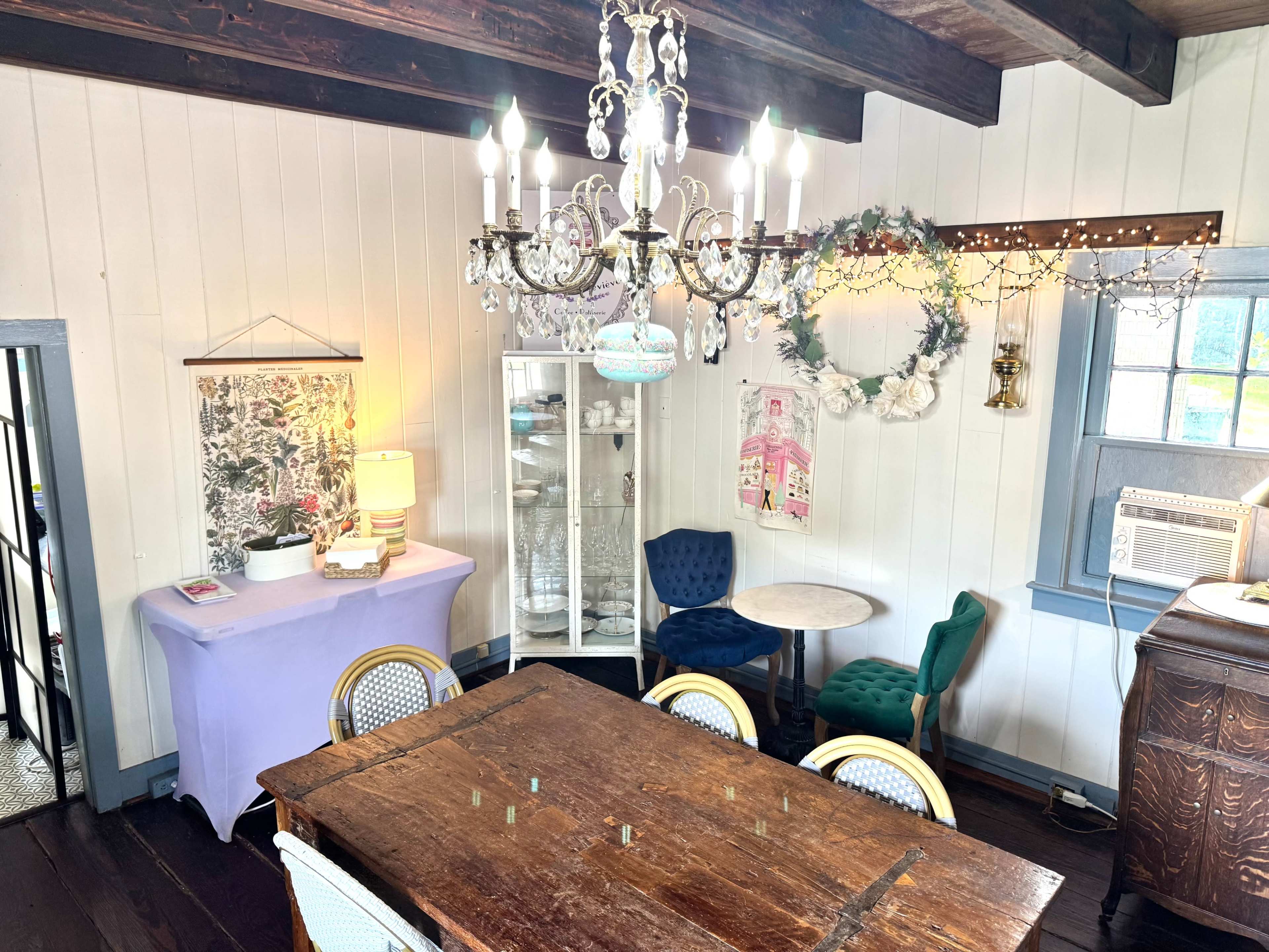 The image shows a cozy dining area with a wooden table, colorful chairs, a chandelier, and a display cabinet against a wall adorned with decor.