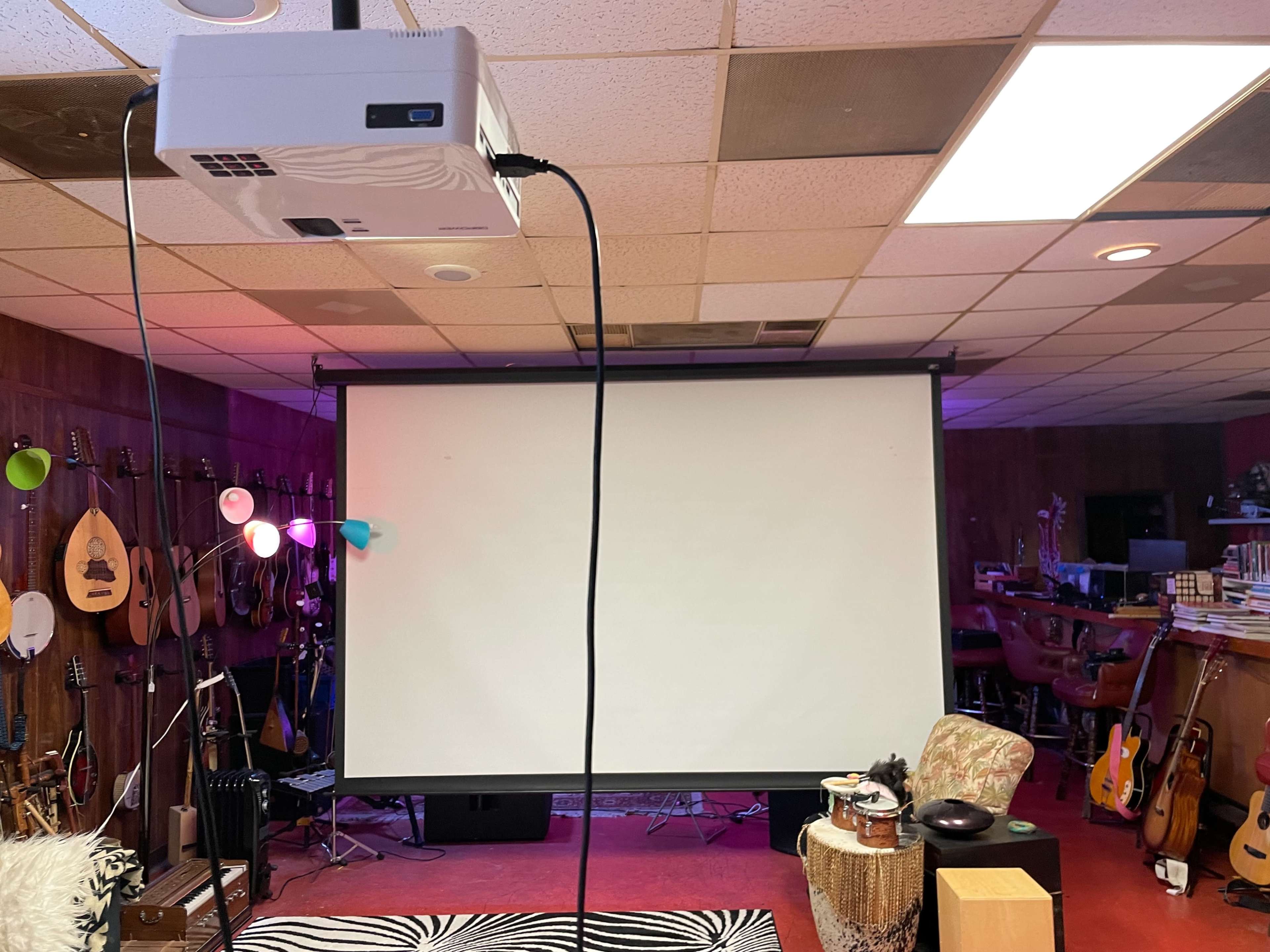 A room with a ceiling-mounted projector pointing at a blank projection screen, surrounded by musical instruments and a seating area.